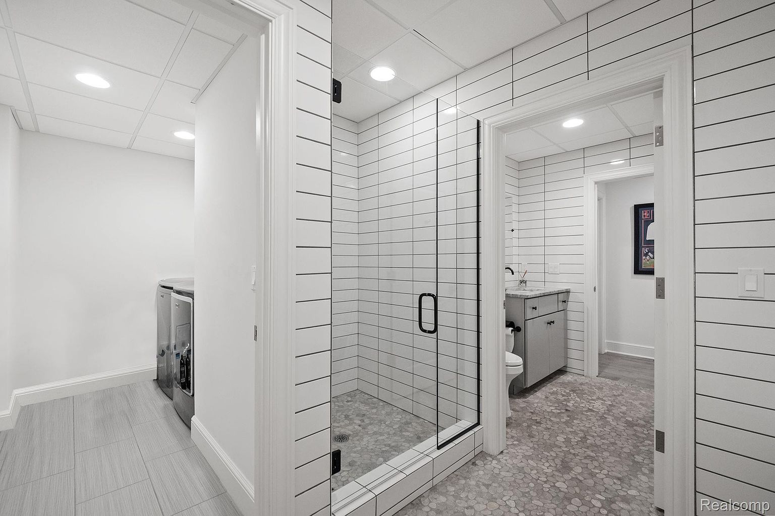 This is a well-lit bathroom featuring a modern design with horizontal white tiles and a pebble-style floor. A glass-enclosed shower is visible, along with a vanity and toilet in a separate area. The space appears clean and stylish, offering a comfortable and contemporary feel.