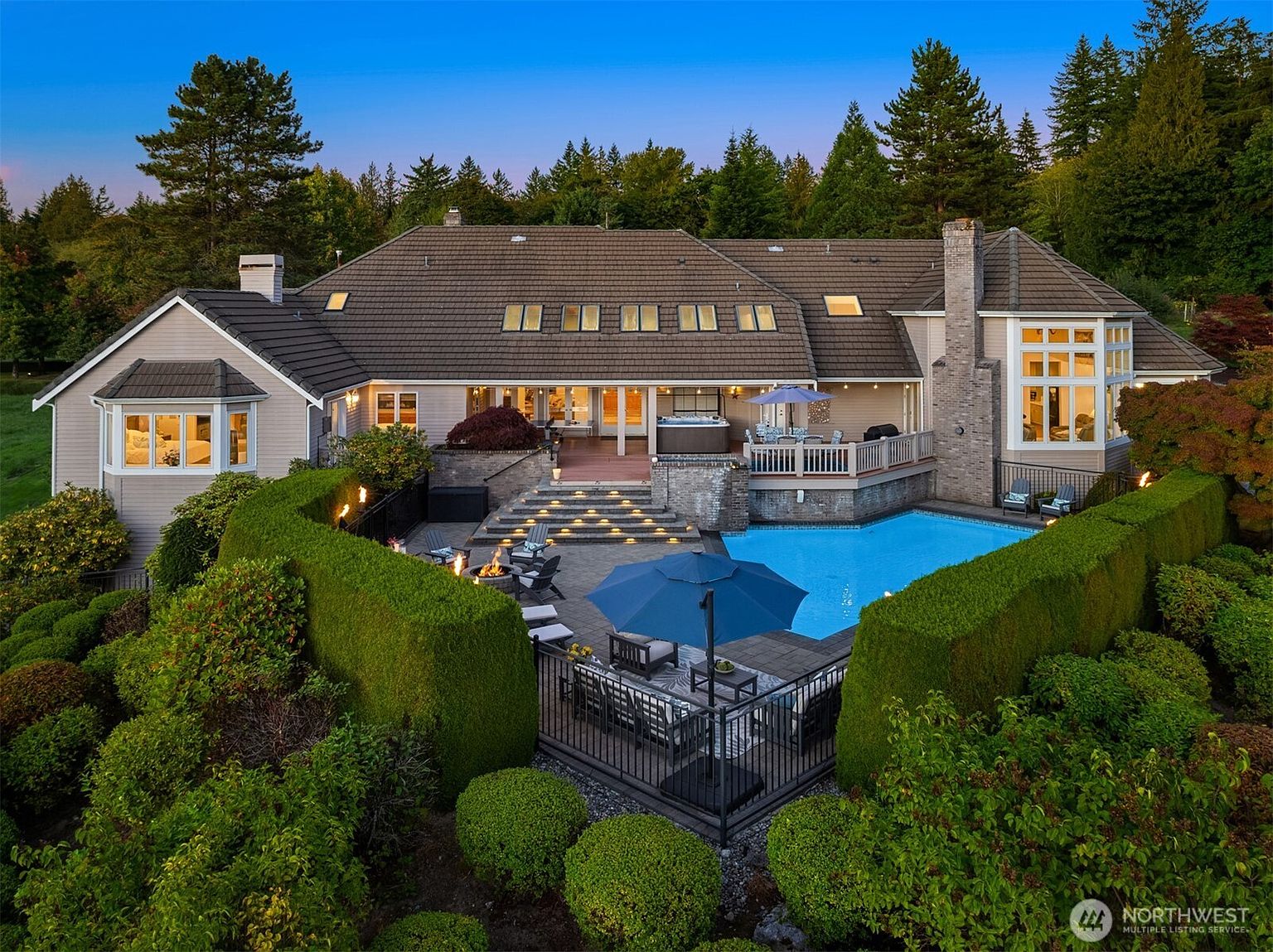 This is an aerial rear view of a luxurious home featuring a large swimming pool, a hot tub on a raised deck, and a spacious patio area with outdoor seating and a fire pit. The property is surrounded by lush greenery and mature trees, creating a private and serene atmosphere. The house itself has a brown roof with multiple skylights and a combination of siding and brick exterior.