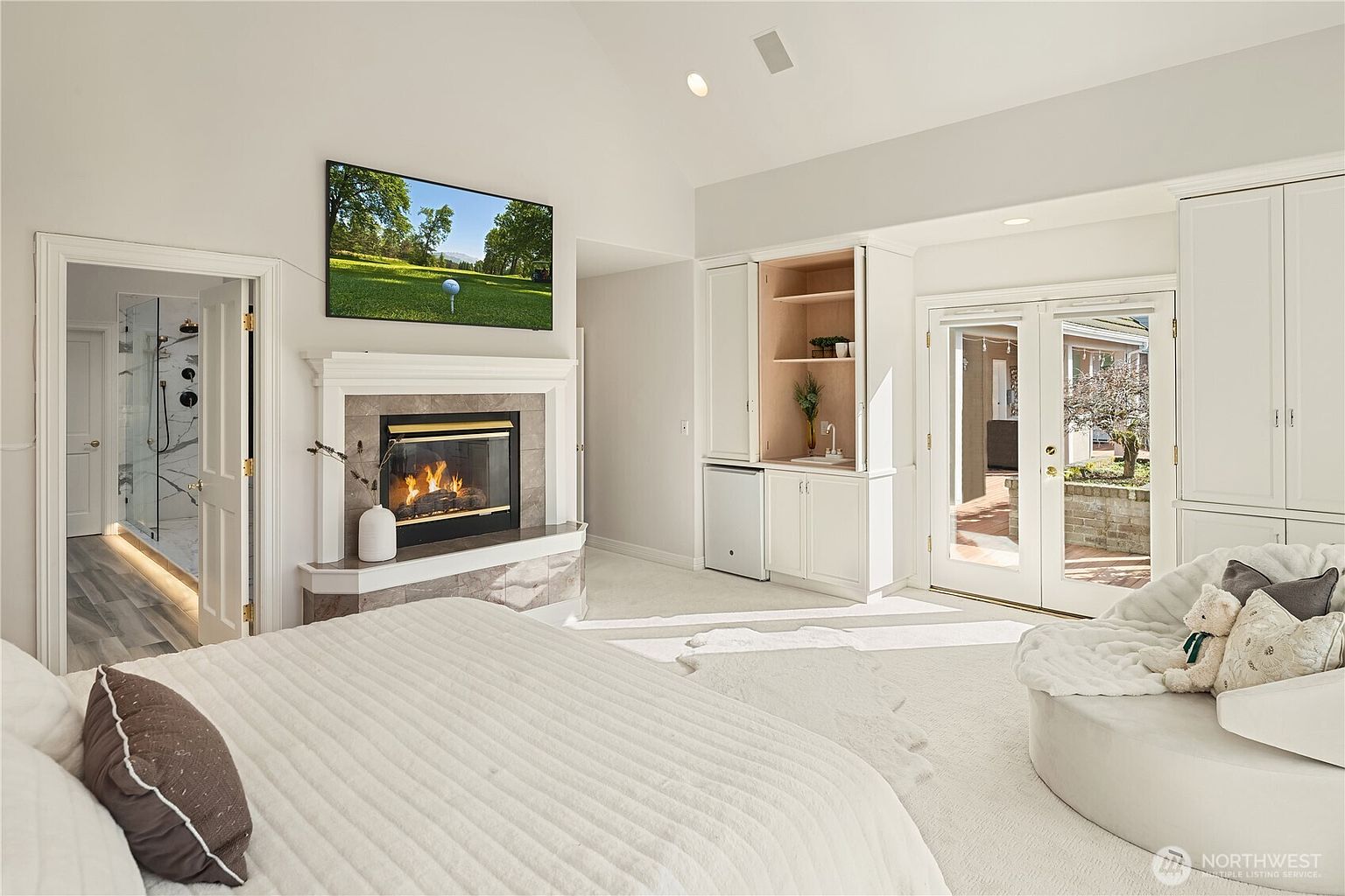 This is a bright and spacious primary bedroom featuring a fireplace with a TV mounted above, built-in cabinetry with a mini-fridge, and French doors leading to an outdoor area. The room is decorated in a neutral color palette with white bedding and carpet, creating a serene and luxurious atmosphere. The perspective is from the foot of the bed, showcasing the room's layout and amenities.