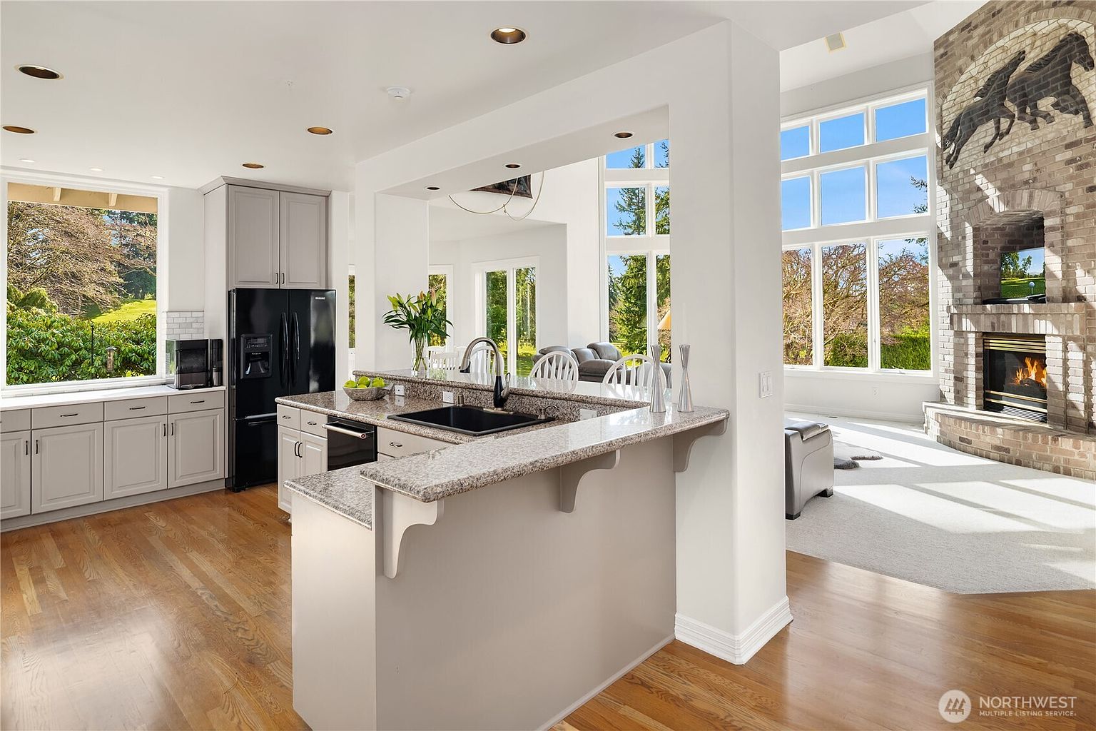 This interior shot showcases a bright and open kitchen that seamlessly transitions into a living area. The kitchen features gray cabinetry, a black refrigerator, and a granite countertop island with a black sink. The living area boasts large windows providing ample natural light and a brick fireplace with a horse art piece above, creating a warm and inviting atmosphere.