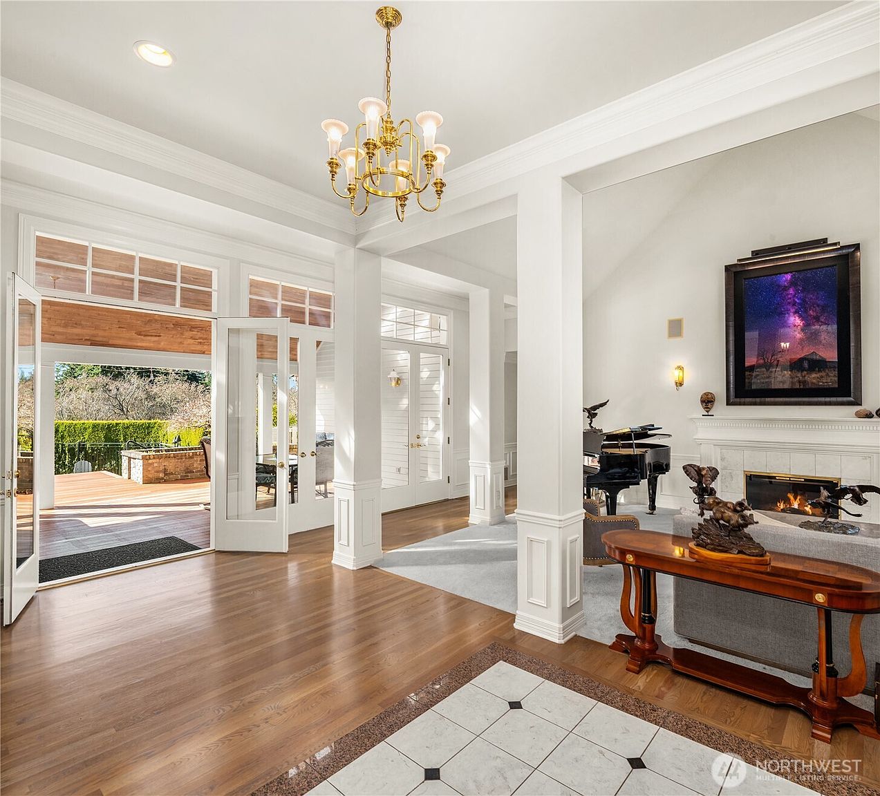 This is an interior shot of a luxurious living room featuring hardwood floors, white columns, and a grand piano. A fireplace with a detailed mantel is visible, along with a large framed artwork above it. The room is well-lit with natural light streaming in from the open doors leading to an outdoor deck, and an elegant chandelier hangs from the ceiling, creating a sophisticated and inviting atmosphere.
