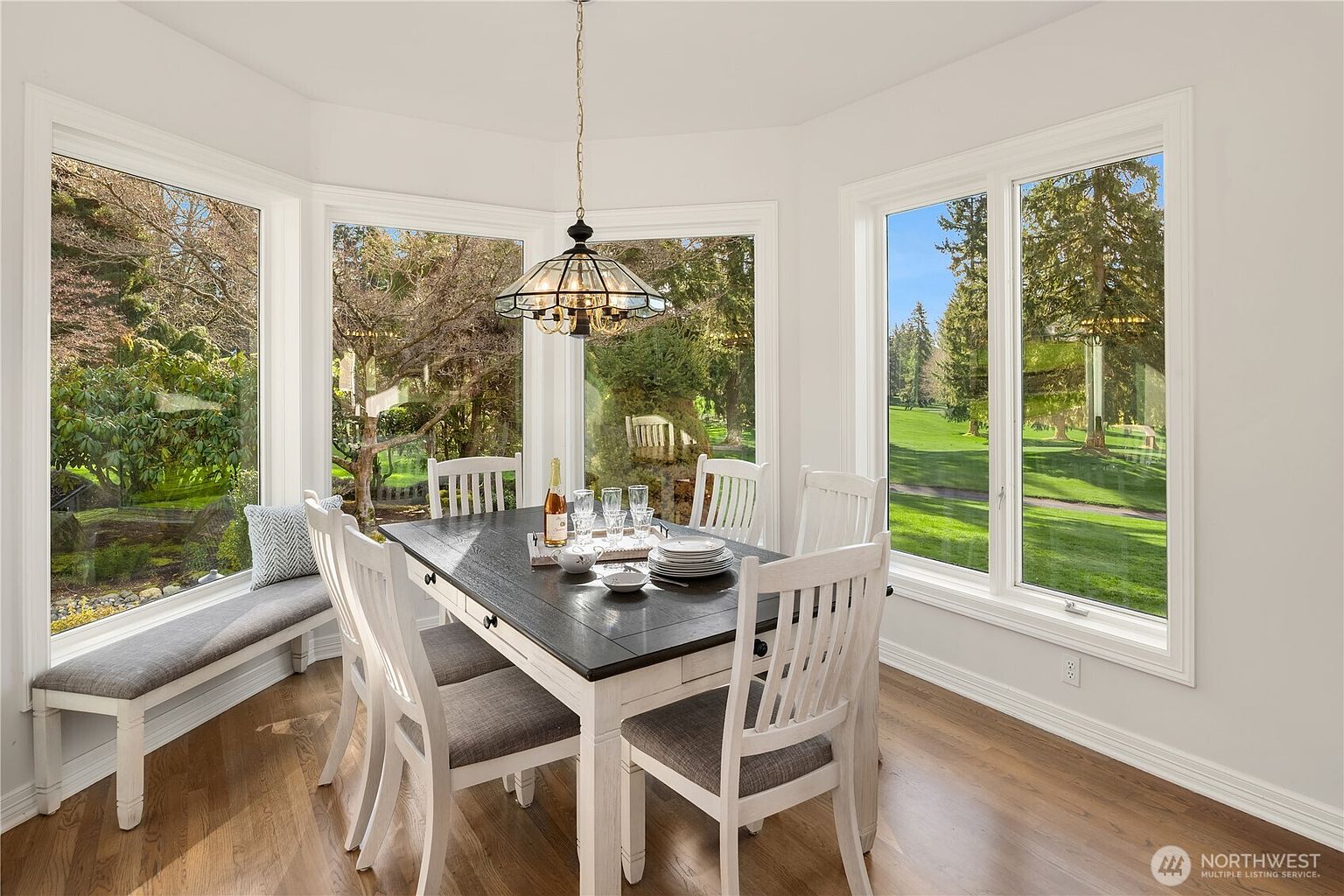 This is an interior shot of a dining room featuring a dark-topped dining table with white legs and matching chairs with gray cushions. A chandelier hangs above the table, and large windows offer views of the surrounding greenery and landscape. A built-in bench seat is visible by one of the windows, creating a cozy and inviting atmosphere.