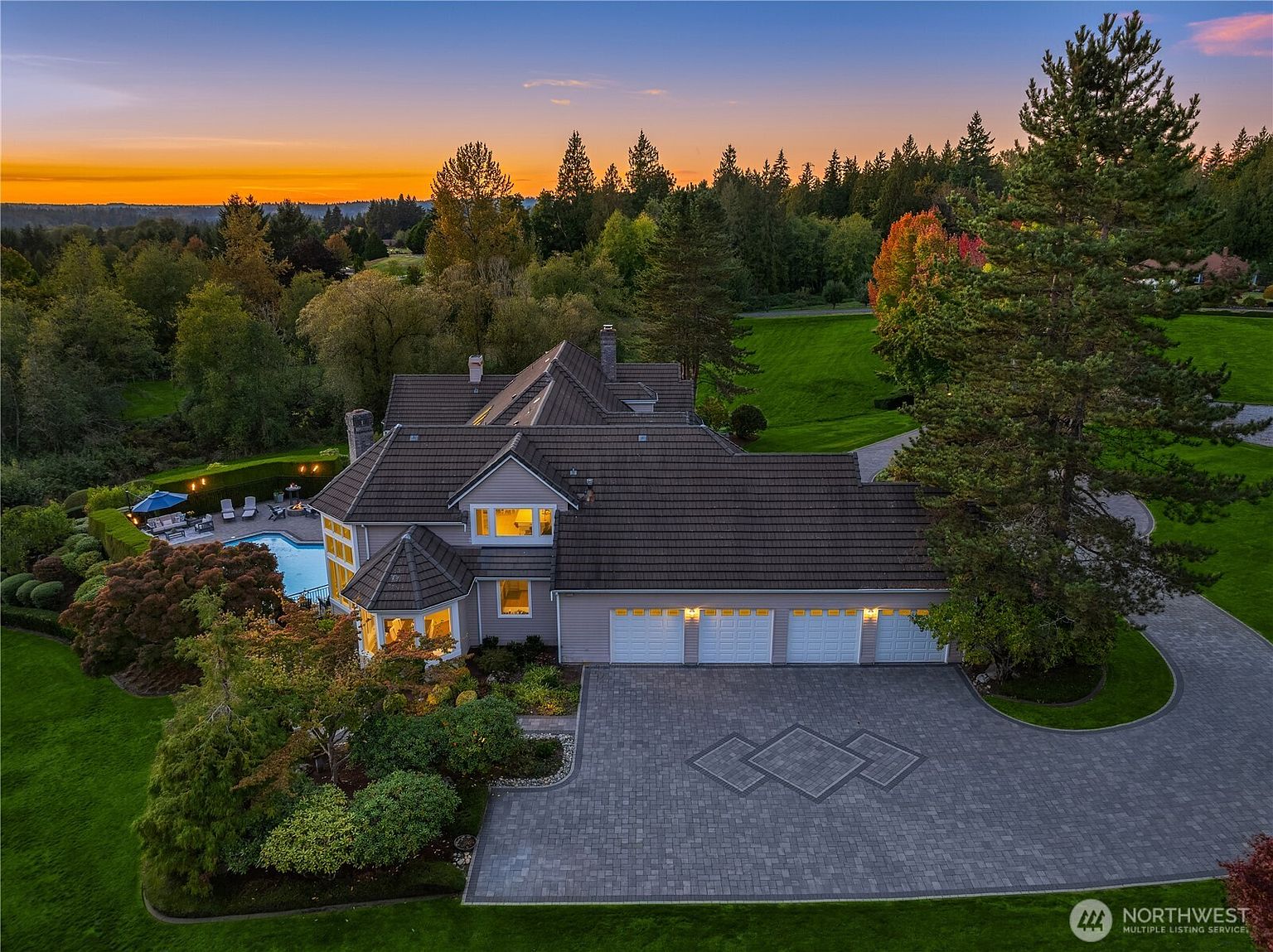 This aerial view showcases a luxurious estate with a sprawling layout, featuring a large house with a multi-car garage, a swimming pool, and meticulously landscaped grounds. The property is surrounded by lush greenery and mature trees, offering privacy and a sense of tranquility. The driveway is paved with intricate brickwork, adding to the overall elegance of the property.