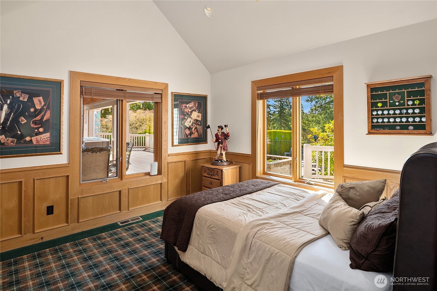 This is a primary bedroom featuring wood paneling, framed artwork, and two large windows that provide natural light and views of the outdoors. The bed is neatly made with a combination of brown and beige bedding, and the room has a cozy, traditional aesthetic. A golf ball display case adds a unique touch.