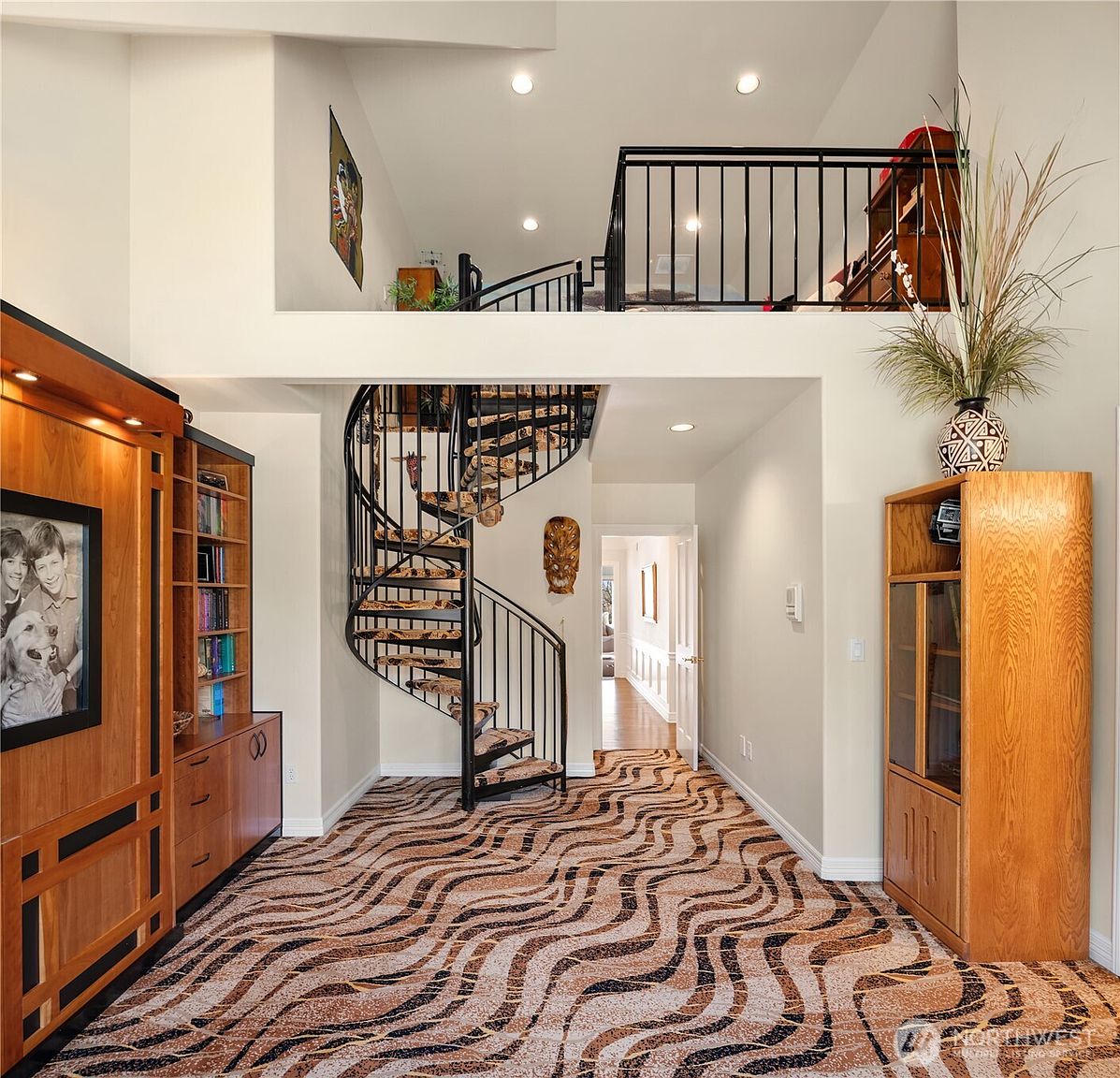 This interior shot showcases a grand hallway featuring a striking spiral staircase with patterned carpeting. The space is well-lit with recessed lighting and natural light, highlighting the unique zebra-striped carpet design. Built-in wooden cabinetry adds a touch of elegance and functionality to the area.