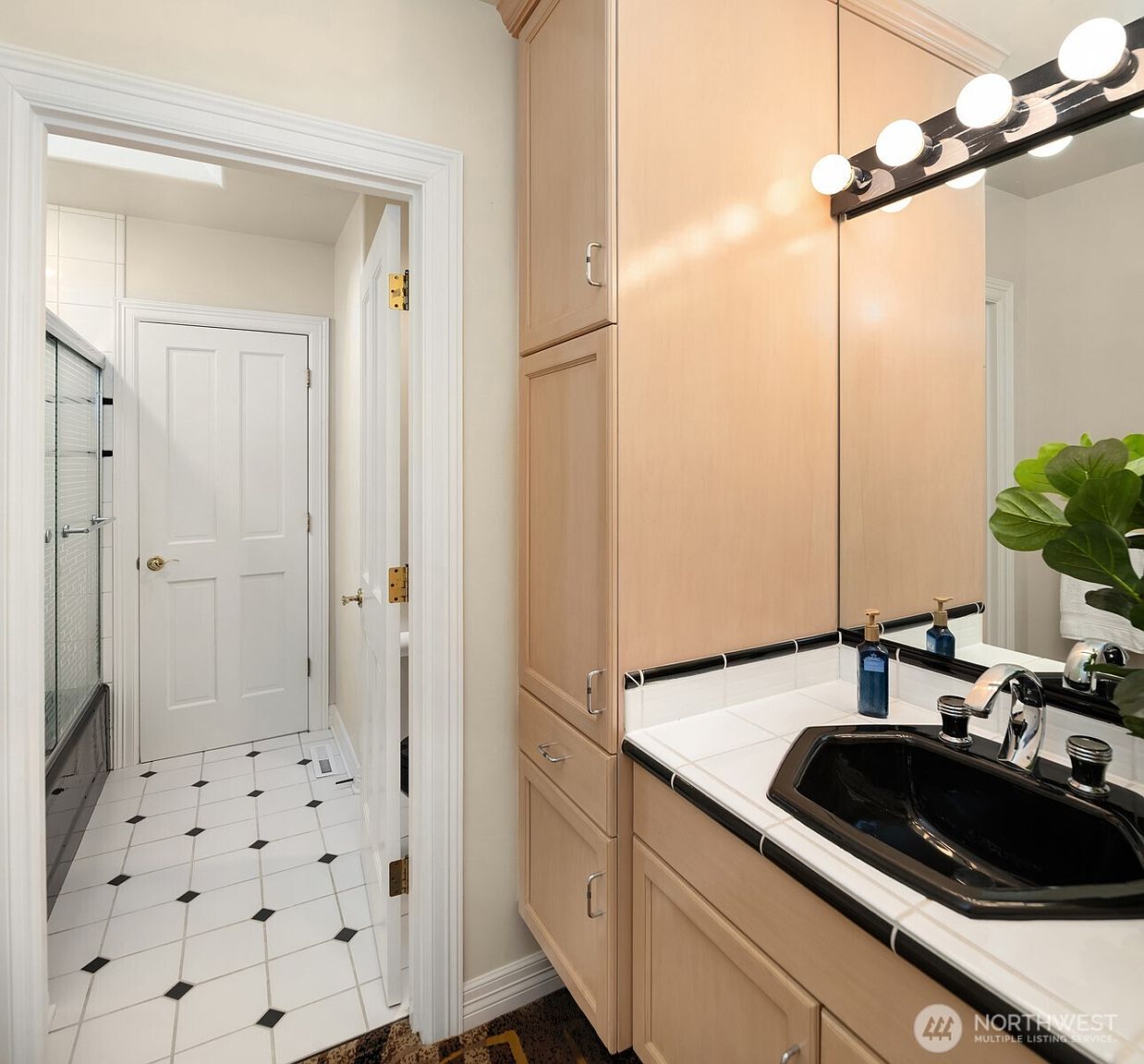 This is a well-lit bathroom featuring a black sink with a chrome faucet set into a white tiled countertop with black trim. The bathroom includes a tall, light wood-colored vanity with storage cabinets and drawers. The floor is tiled in a white and black checkerboard pattern, and a doorway leads to a shower and toilet area.