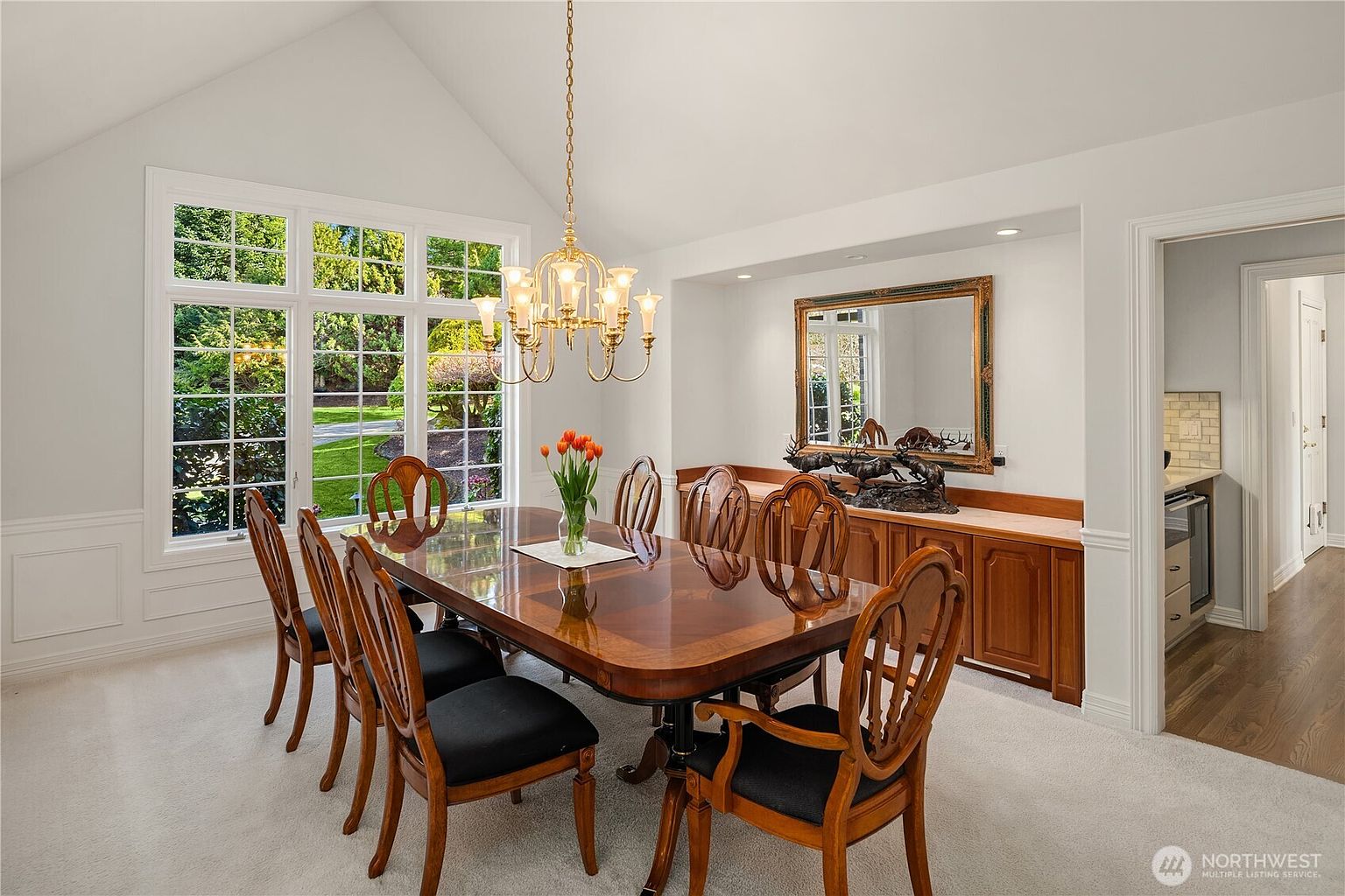This is an interior shot of a dining room featuring a large wooden dining table with black cushioned chairs. A gold chandelier hangs above the table, and a large window provides natural light and a view of the exterior greenery. A wooden cabinet with a decorative mirror sits against the wall, adding to the room's traditional and elegant style.