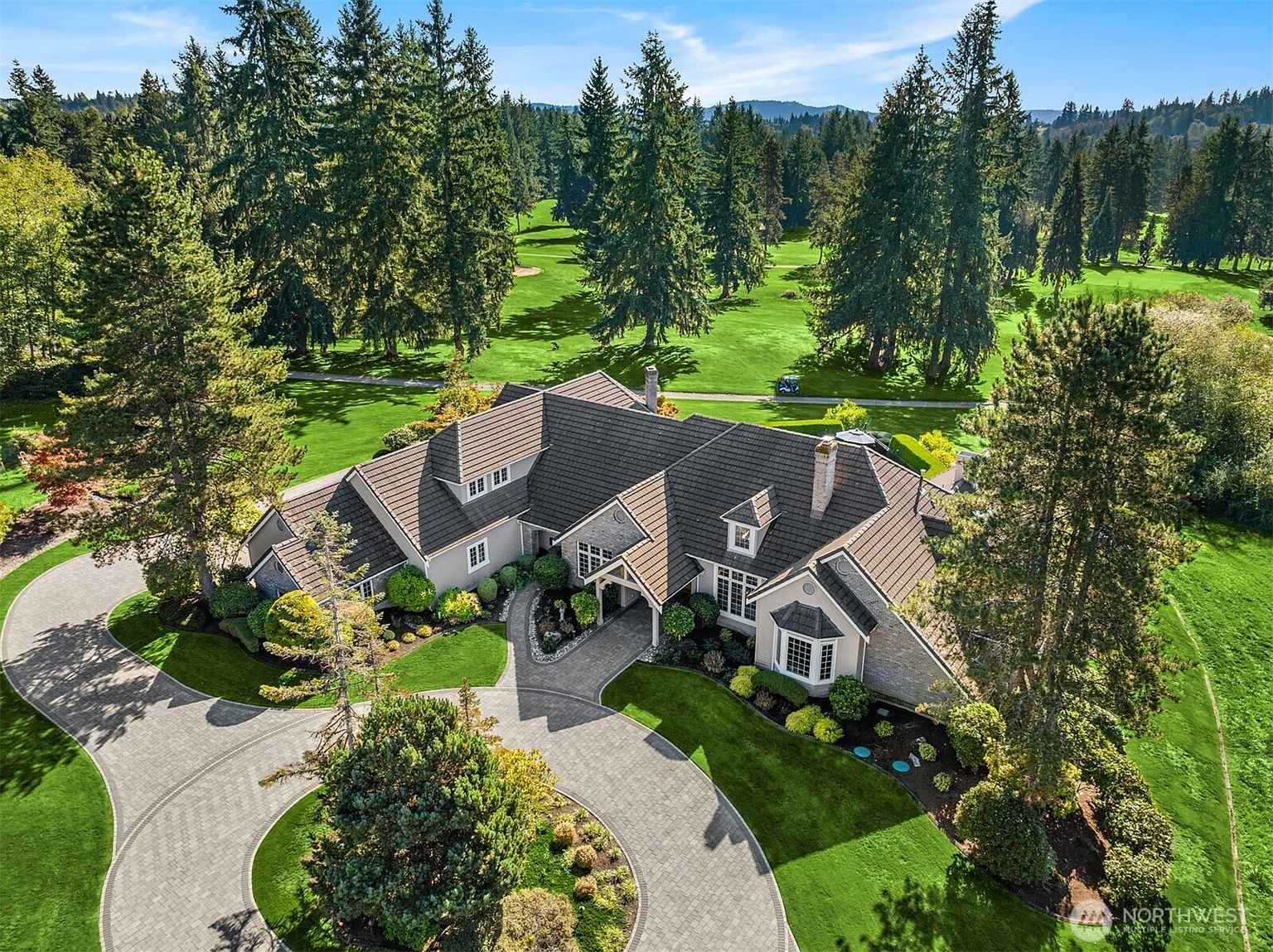This aerial view showcases a luxurious home with a meticulously landscaped circular driveway and lush green lawns. The house features a complex roofline with multiple gables and dormers, complemented by mature trees and a golf course in the background. The overall impression is one of elegance and privacy, highlighting the property's expansive grounds and desirable location.