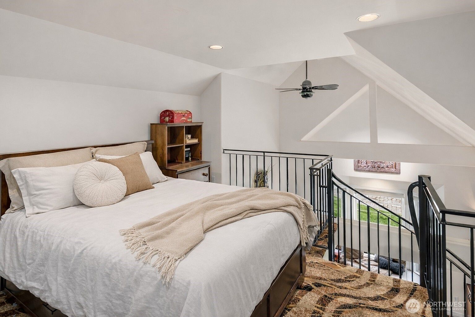 This is a cozy primary bedroom featuring a bed with white linens and decorative pillows, a wooden headboard, and a small wooden cabinet. The room has an attic-like feel with angled ceilings and a view of a staircase with black railings. The overall impression is warm and inviting.