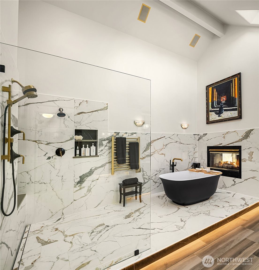 This is a luxurious primary bathroom featuring marble-like tile throughout, a glass-enclosed shower with gold fixtures, and a freestanding black bathtub. A modern fireplace adds warmth and ambiance, while a piece of art hangs above the tub. The space is well-lit and exudes a spa-like atmosphere.
