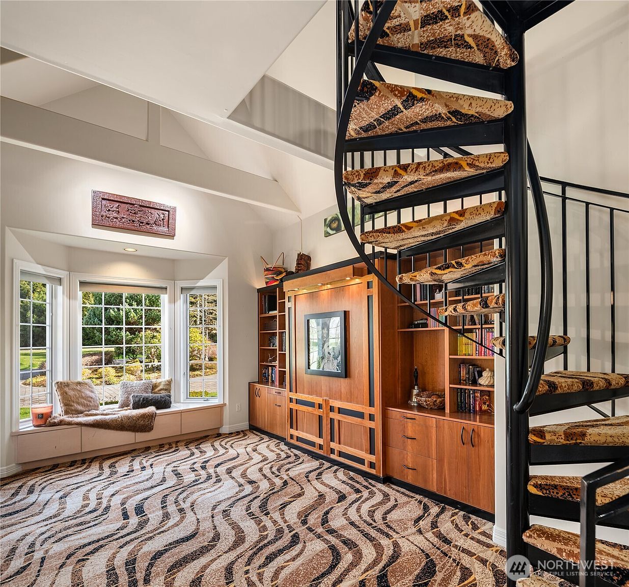 This interior shot showcases a unique living room with a spiral staircase and a built-in entertainment center. A bay window provides ample natural light and a view of the outdoors, while the patterned carpet adds a touch of character. The room's design blends functionality with distinctive architectural elements.