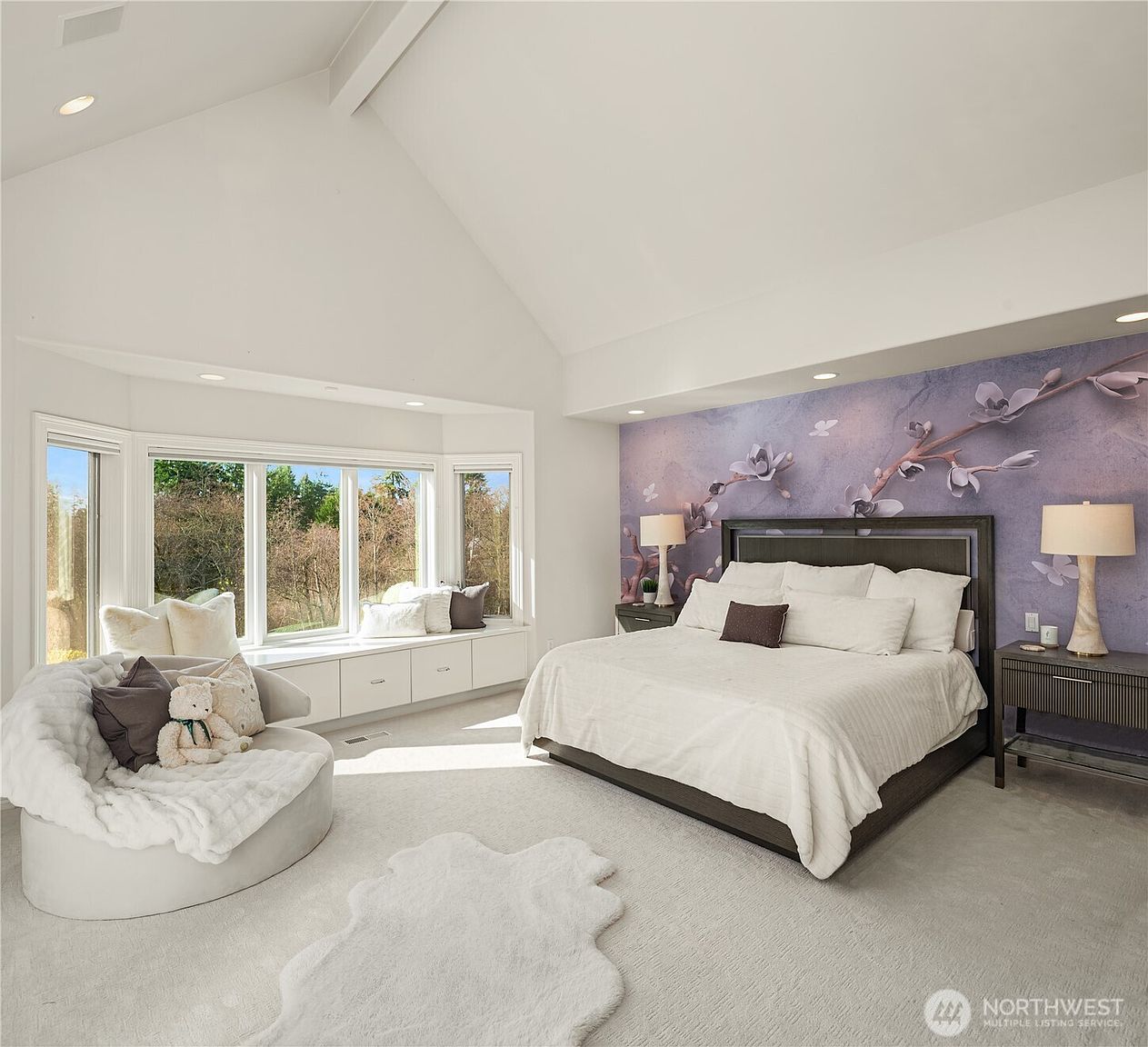 This is a bright and spacious primary bedroom featuring a large bed with white linens, a statement wall with floral wallpaper, and a cozy seating area by a bay window. The room is carpeted in a light color, and the overall design is elegant and serene, creating a relaxing atmosphere. The perspective is from the doorway, showcasing the entire room.
