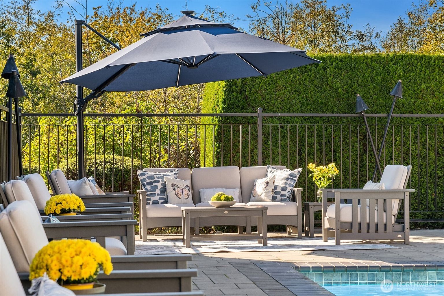 This image showcases a beautifully arranged outdoor patio area, perfect for relaxation and entertaining. The patio features comfortable seating, including a sofa and chairs, arranged around a coffee table, all shaded by a large umbrella. The setting is enhanced by decorative pillows, floral arrangements, and tiki torches, creating a welcoming and stylish outdoor living space.