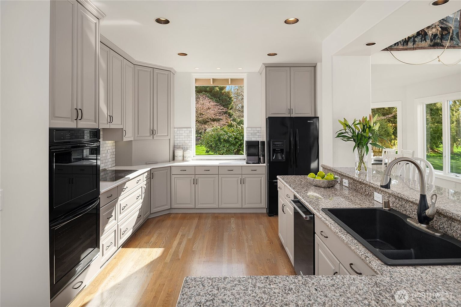 This is a well-lit kitchen featuring light gray cabinetry with black hardware, stainless steel appliances, and granite countertops. A large window provides natural light and a view of the outdoors. The kitchen has a modern and clean aesthetic, with hardwood floors adding warmth to the space.