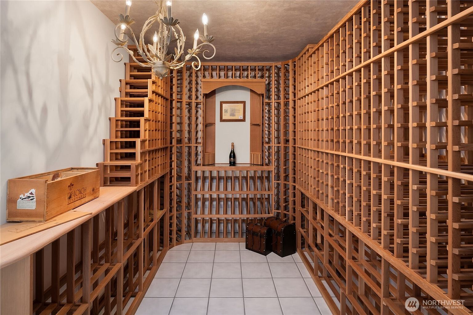 This is a well-organized wine cellar featuring custom wooden racking that fills the room from floor to ceiling. A decorative chandelier hangs above, and the room is lit to showcase the wine collection. The floor is tiled, and there are a few decorative items such as wooden chests and a wine box, adding to the ambiance.