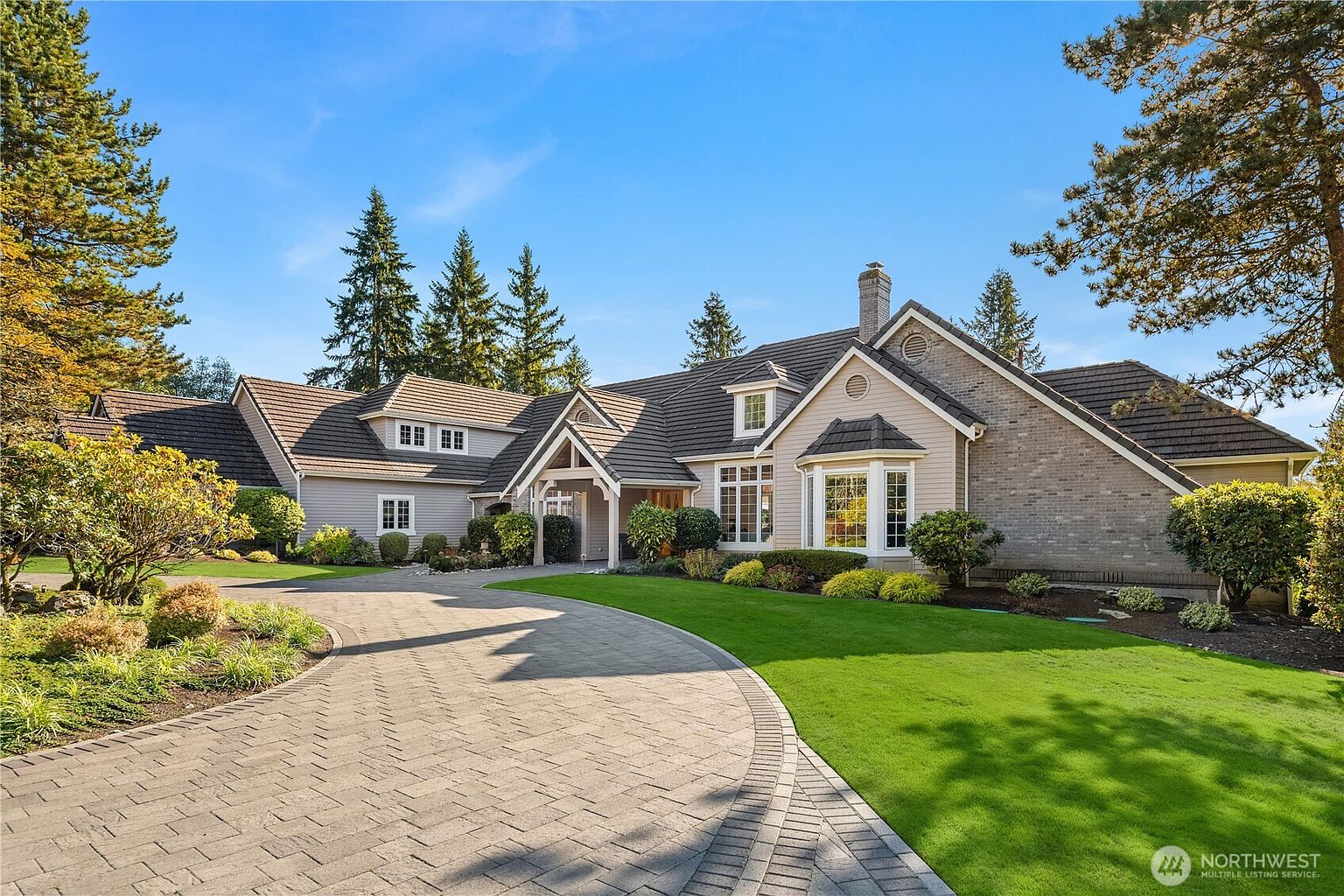 This is a front exterior view of a luxurious single-family home. The house features a well-manicured lawn, a circular driveway paved with bricks, and a combination of light-colored siding and brick accents. The roof is dark and multi-gabled, with several dormer windows, creating an elegant and inviting curb appeal.