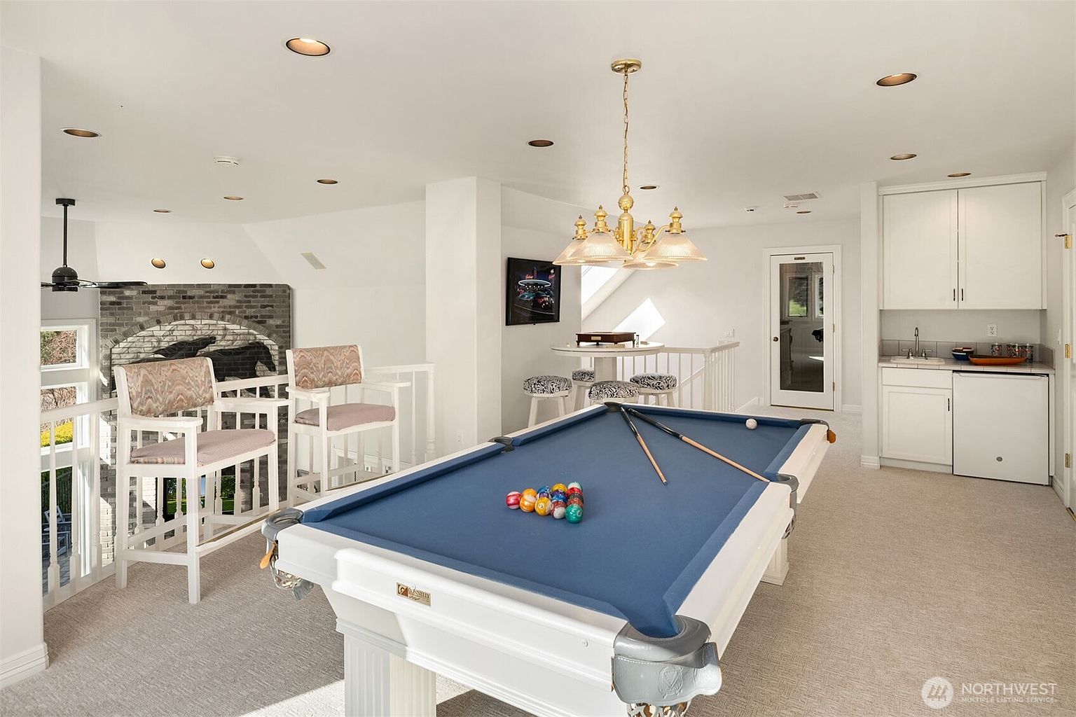 This interior shot showcases a well-lit living room featuring a pool table as a central element. The room is decorated in a classic style with white walls, a chandelier, and comfortable seating. A bar area with cabinets and a sink is visible in the background, adding to the entertainment space.