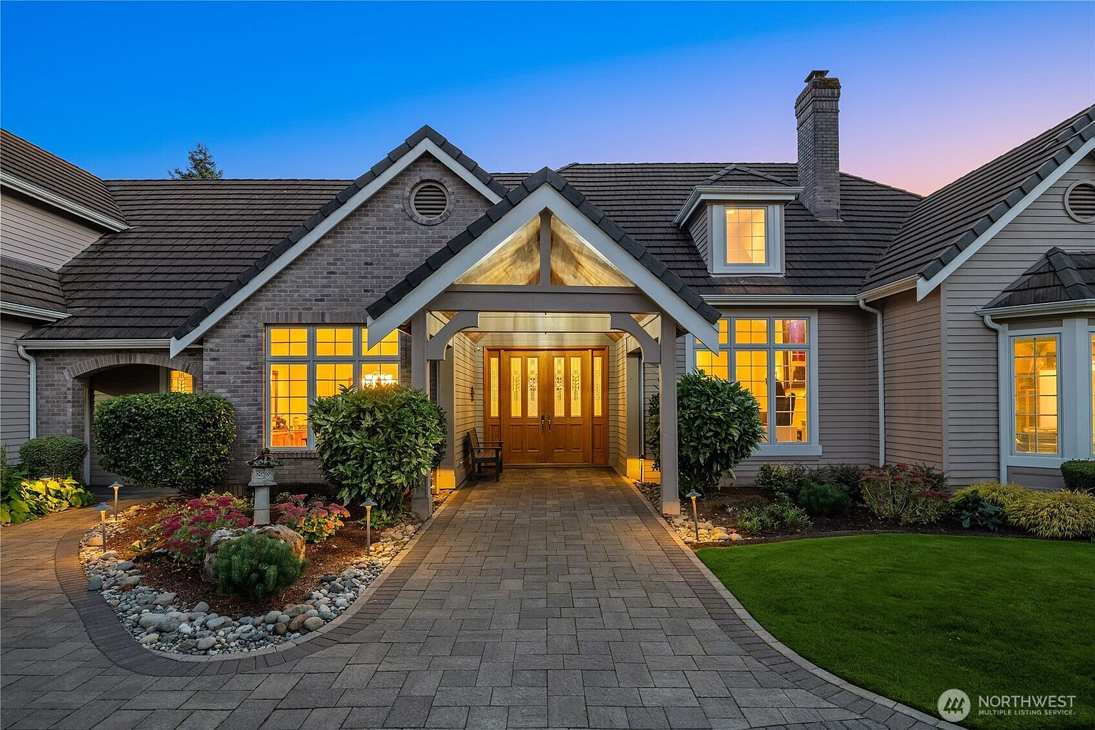 The image showcases the entryway of a well-maintained home at dusk. A paved walkway leads to a covered porch with a wooden double door, flanked by manicured bushes and flower beds. Warm light emanates from the windows, creating an inviting and elegant ambiance.