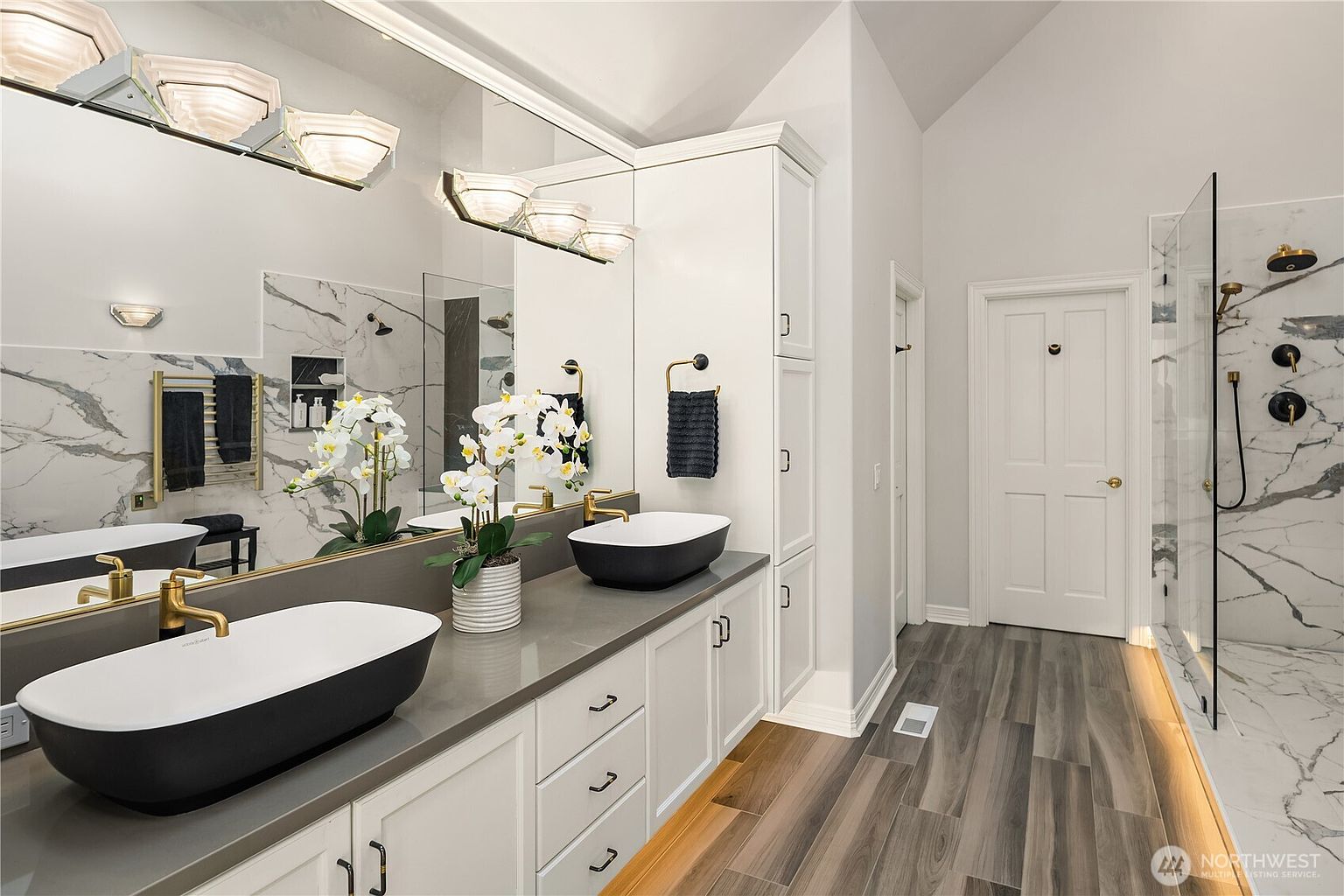 This is a primary bathroom featuring a double vanity with black and white vessel sinks, gold fixtures, and white cabinetry. A large mirror reflects the sinks and lighting, while a glass-enclosed shower with marble-like tile is visible on the right. The flooring is wood-look tile, and the overall style is modern and luxurious.