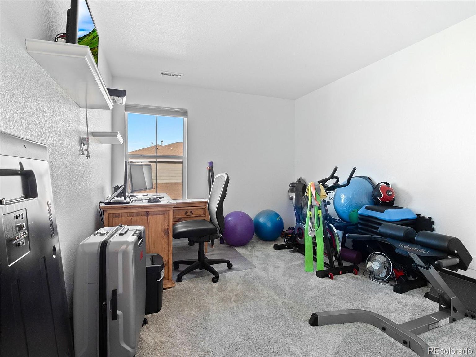 This is an interior shot of a home gym. The room features a desk with a computer, a rolling office chair, a suitcase, and various exercise equipment including an exercise bike, stability balls, and a weight bench. The walls are painted white, and the floor is carpeted, creating a functional and versatile space.