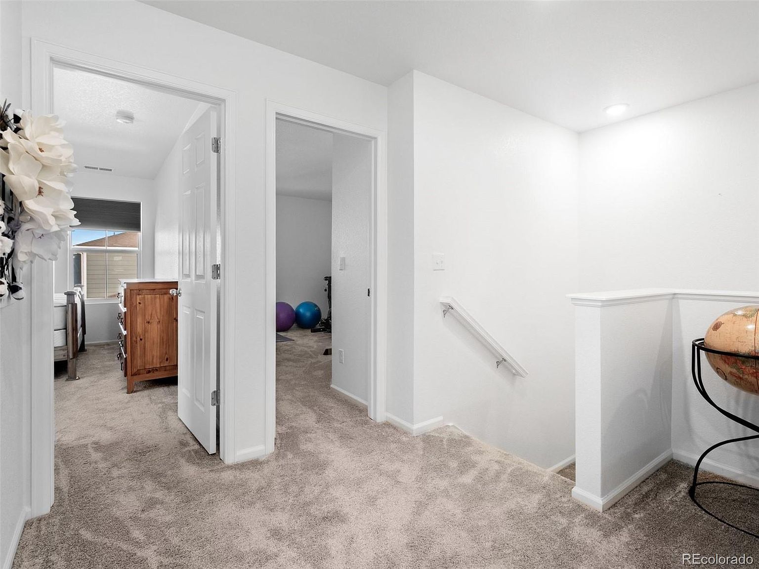 This interior shot showcases a well-lit hallway with neutral-toned carpet and white walls, creating a bright and airy atmosphere. Two doorways lead to adjacent rooms, one revealing a bedroom and the other a space with exercise equipment. A staircase with a white banister descends to another level, adding depth and visual interest to the space.