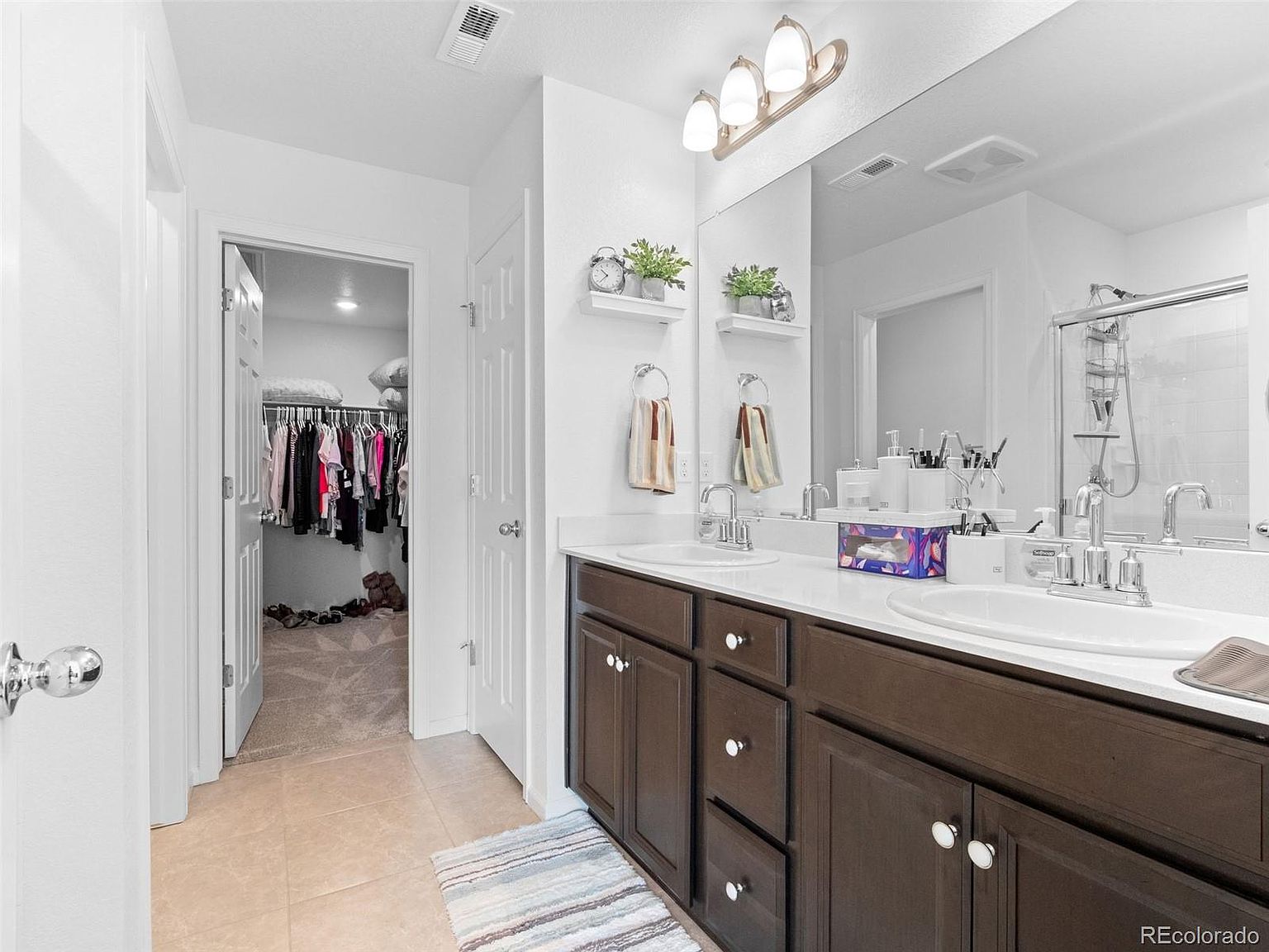 This is a well-lit primary bathroom featuring a double vanity with dark wood cabinets and white countertops. A large mirror hangs above the sinks, reflecting the bright lighting and adding to the spacious feel. An open walk-in closet is visible through a doorway, suggesting ample storage space.