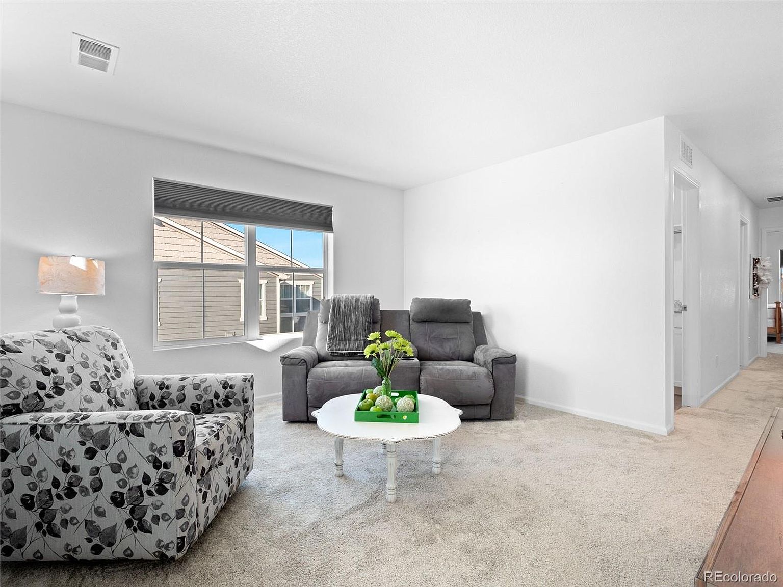 This is a bright and inviting living room featuring neutral-toned carpet and white walls. The room is furnished with a floral armchair, a gray loveseat, and a white round coffee table adorned with a green tray and floral arrangement. A window with blinds provides natural light, and a hallway is visible to the right, creating an open and airy feel.