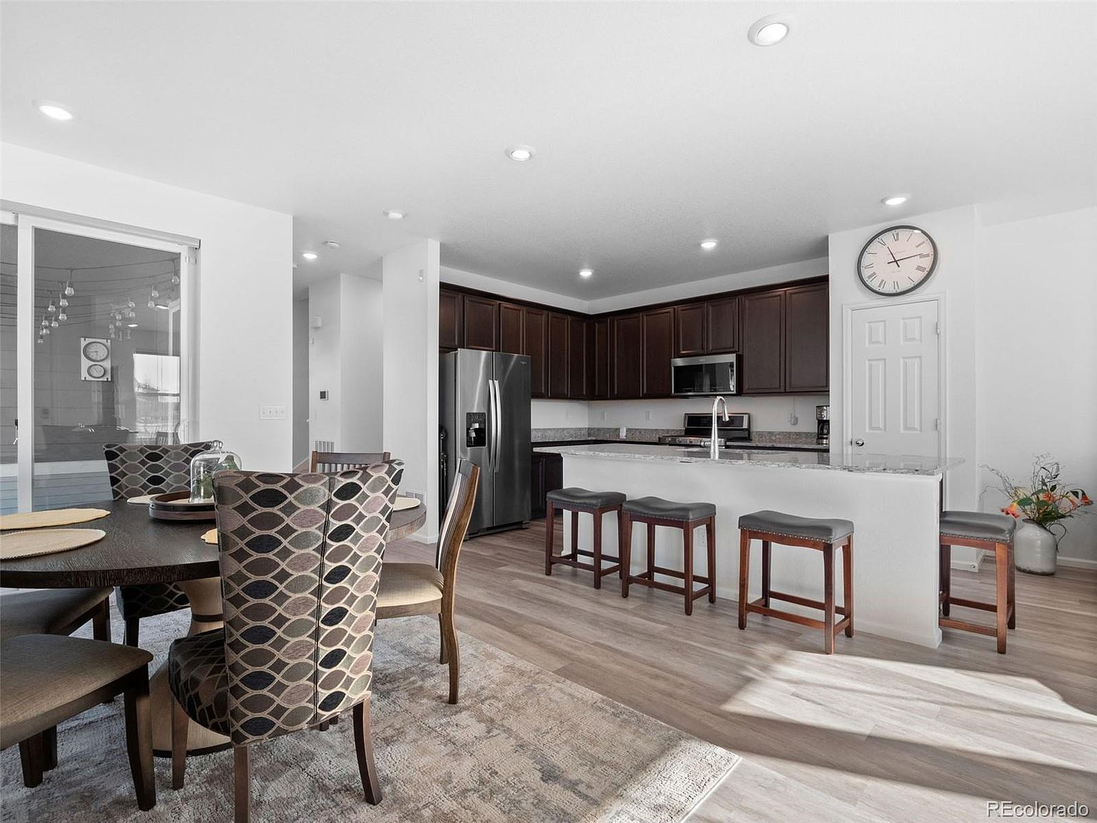 This is an interior shot of a modern kitchen and dining area. The kitchen features dark wood cabinetry, stainless steel appliances, and a breakfast bar with seating. The dining area includes a round table with upholstered chairs, set on a neutral-toned rug, creating a cohesive and inviting space.