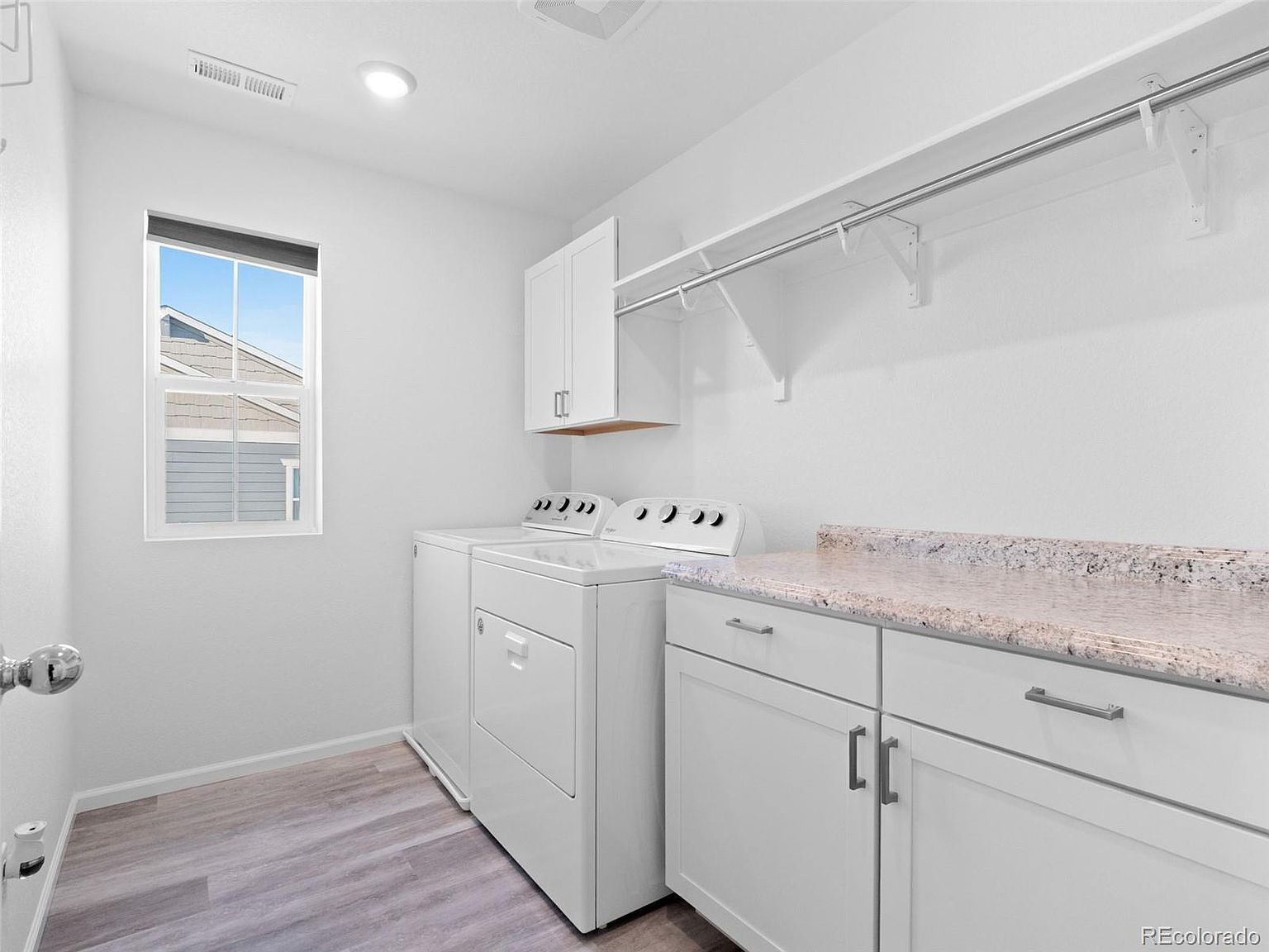 This is a bright and clean laundry room featuring a white washer and dryer set, white cabinetry with granite countertops, and a hanging rod for clothes. A window provides natural light, and the light wood-look flooring adds a touch of warmth. The room appears well-organized and functional.
