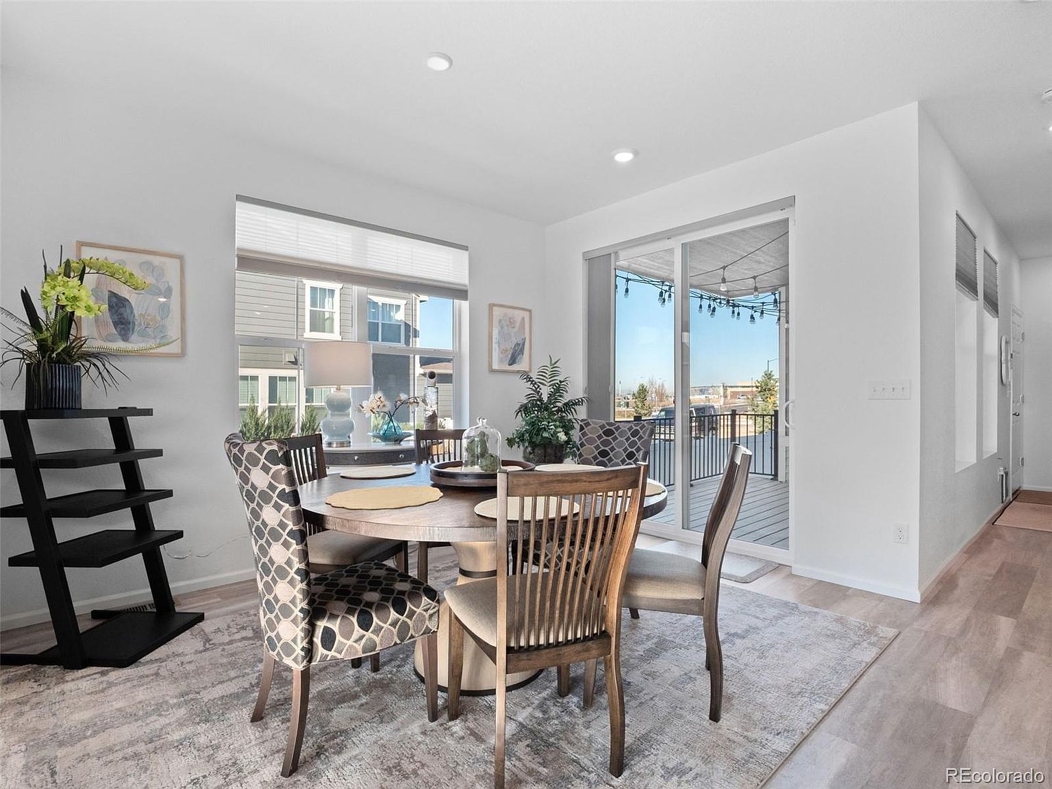 This is an interior shot of a dining room featuring a round wooden table with four chairs, a decorative rug, and a black shelving unit. Natural light floods the room through a large window and sliding glass doors leading to a balcony, creating a bright and inviting atmosphere. The room's neutral color palette and contemporary furnishings contribute to a stylish and comfortable dining space.