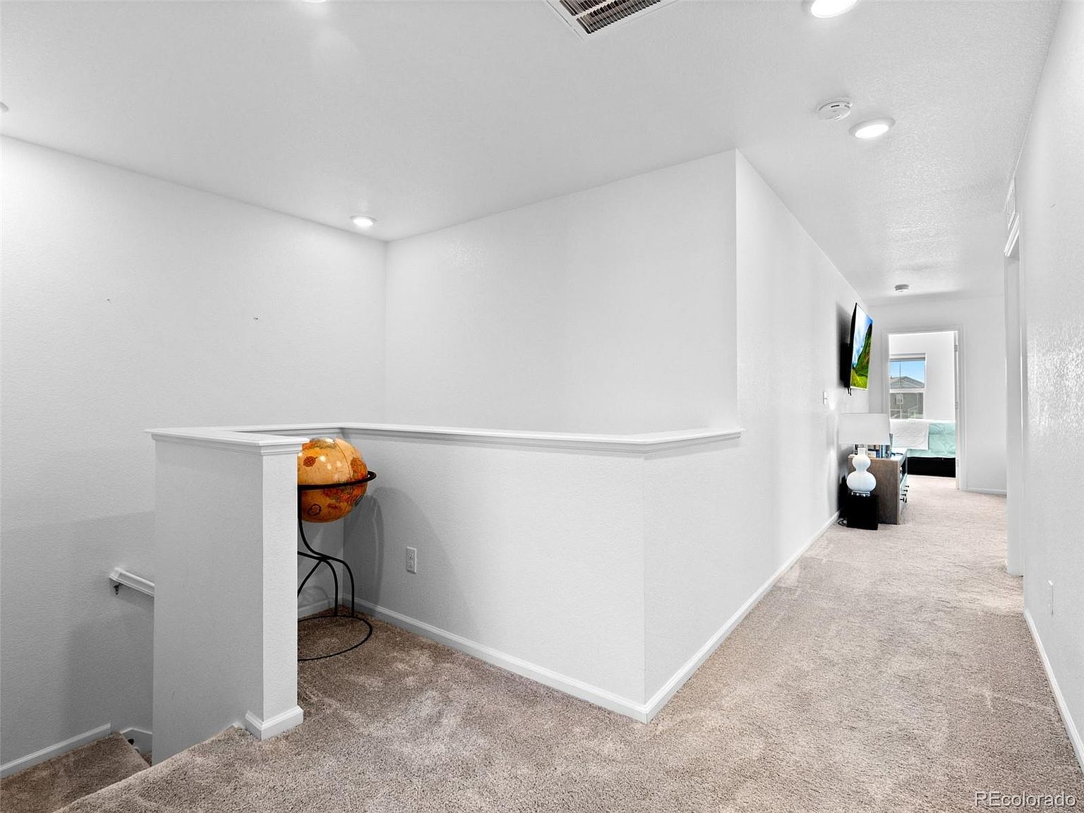 This is an interior shot of a hallway and stairs. The hallway is carpeted in a neutral tone and features white walls and trim. A decorative globe sits on a stand near the stairs, adding a touch of character. The hallway leads to a bedroom, visible in the background, suggesting a well-maintained and inviting home.