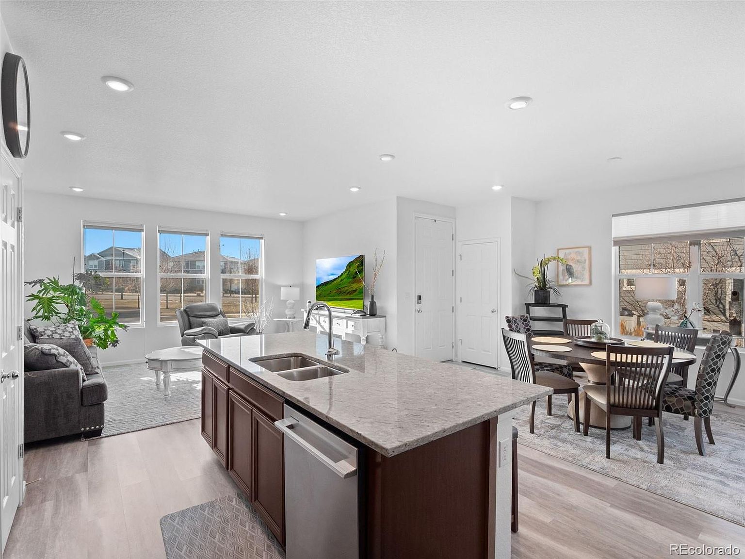 This is an open-concept kitchen and living area featuring a large kitchen island with a sink and stainless steel dishwasher. The kitchen flows into a dining area with a round table and chairs, and further into a living room with a gray sofa and arm chair. The space is well-lit with natural light from the windows and recessed lighting, creating a bright and inviting atmosphere.