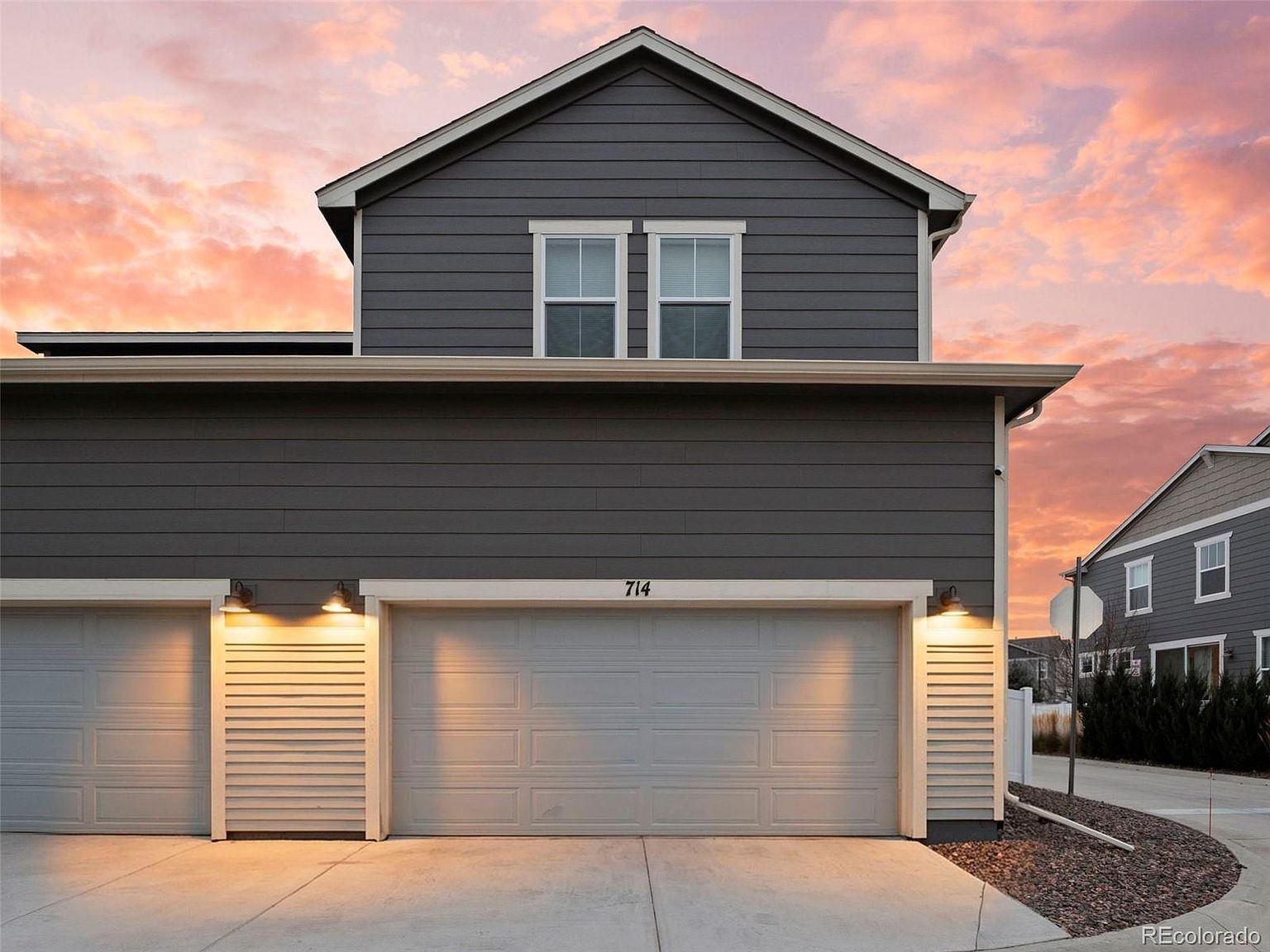 The image showcases the front exterior of a two-story home, focusing on the garage area. The house features gray siding with white trim, and the garage has two doors, one of which has the number '714' above it. The driveway is concrete, and the sky is filled with a beautiful sunset, creating a warm and inviting atmosphere.