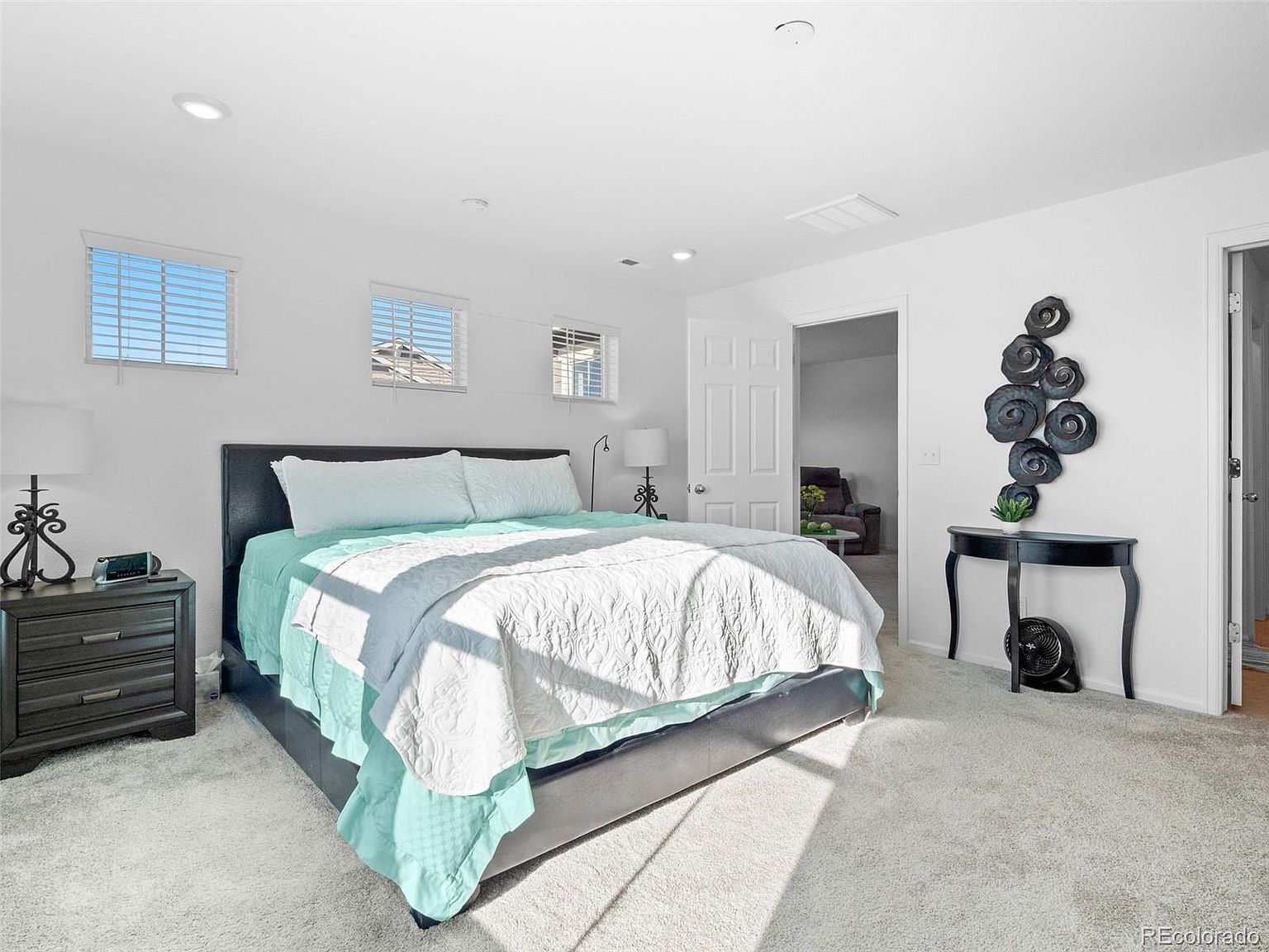 This is a bright and airy primary bedroom featuring a large bed with a light blue comforter and white quilt. The room has white walls, three windows with blinds, and light-colored carpeting. A dark wood nightstand sits beside the bed, and a decorative table with a modern art piece adds a touch of elegance.