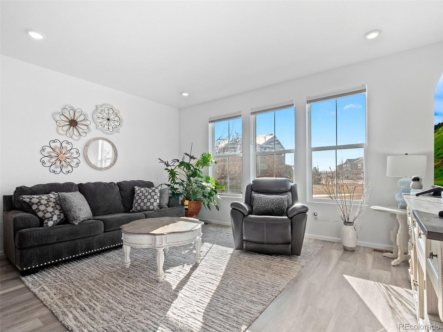 This is a bright and airy living room featuring a gray sofa with decorative pillows, a round coffee table, and a comfortable-looking gray recliner. Large windows provide ample natural light, and the neutral color palette creates a calming atmosphere. The room is decorated with wall art and a potted plant, adding a touch of personality.