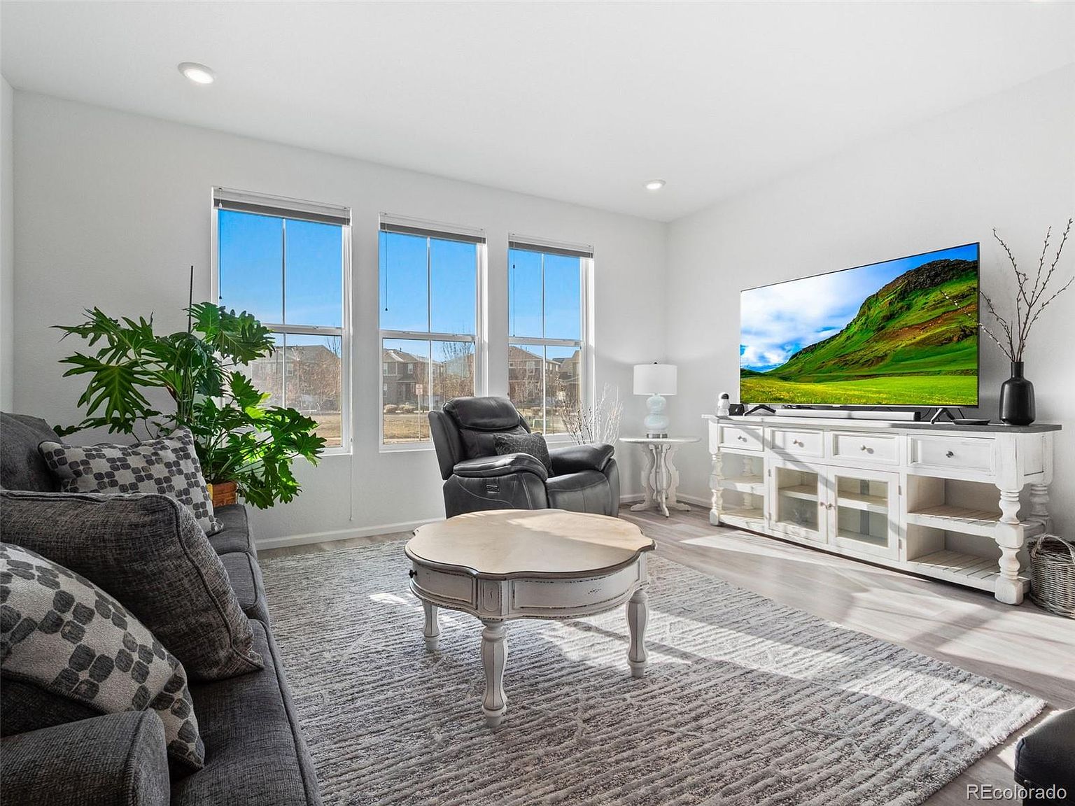 This is a bright and airy living room featuring a gray sofa with patterned pillows, a round coffee table, and a gray recliner. Large windows provide ample natural light, and a white entertainment center houses a large television. The room is decorated in neutral tones with a modern aesthetic, creating a welcoming and comfortable atmosphere.