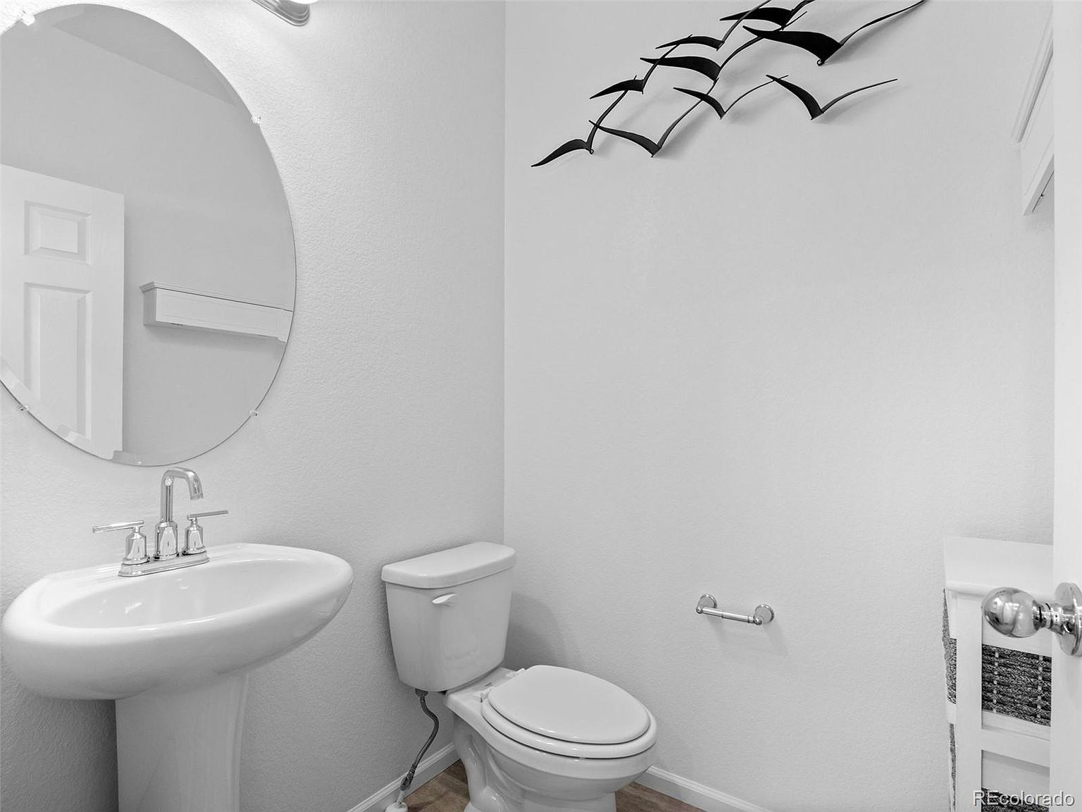 This is a well-lit guest bathroom featuring a pedestal sink with a chrome faucet, a round mirror, and a standard toilet. The walls are painted in a neutral color, and a decorative metal bird sculpture adds a touch of modern art. The overall impression is clean and functional, suitable for a guest or secondary bathroom.