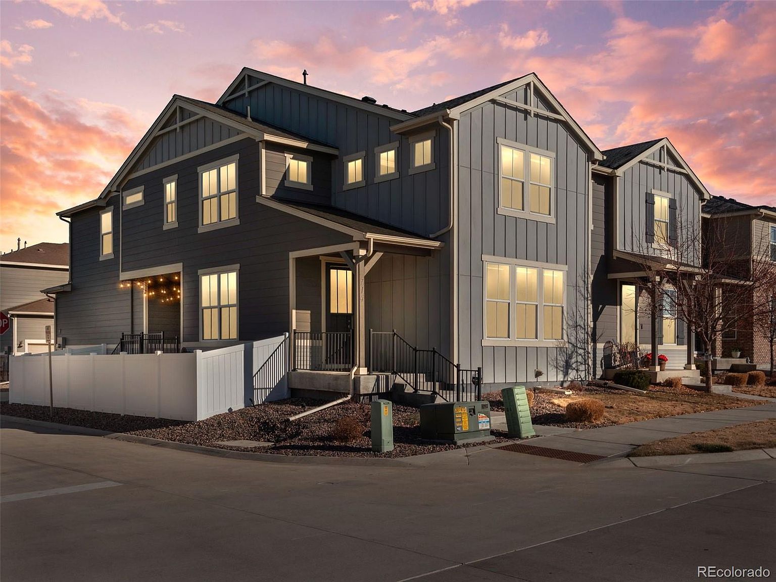 This is a front view of a modern two-story townhome with a gray exterior and white trim. The home features multiple windows that allow ample natural light, a small front porch with black railings, and well-maintained landscaping. The property is situated on a corner lot with a paved street in front, creating a welcoming curb appeal.