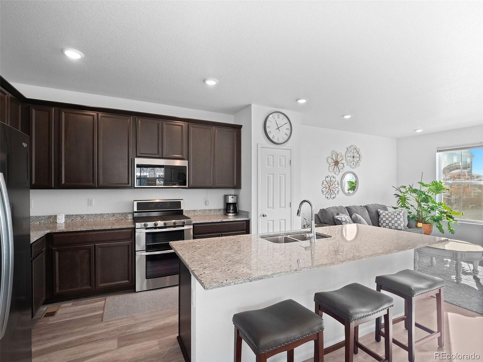 This is a well-lit kitchen featuring dark wood cabinetry, granite countertops, and stainless steel appliances. A large island with bar seating anchors the space, providing a gathering spot. The kitchen seamlessly flows into the living area, creating an open and inviting atmosphere.