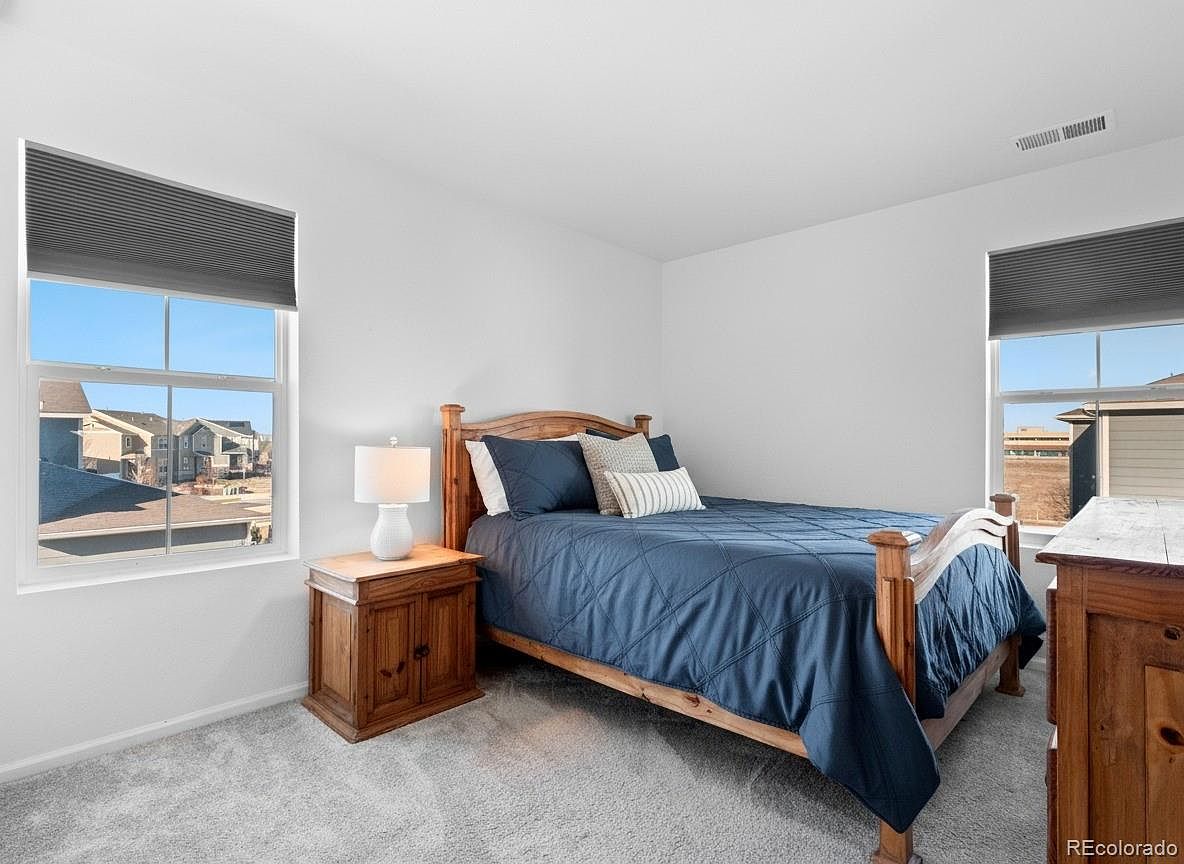 This is a cozy bedroom featuring a wooden bed with blue bedding and decorative pillows. A wooden nightstand with a lamp sits beside the bed, and a window provides natural light. The room has a neutral color palette and a carpeted floor, creating a comfortable and inviting atmosphere.