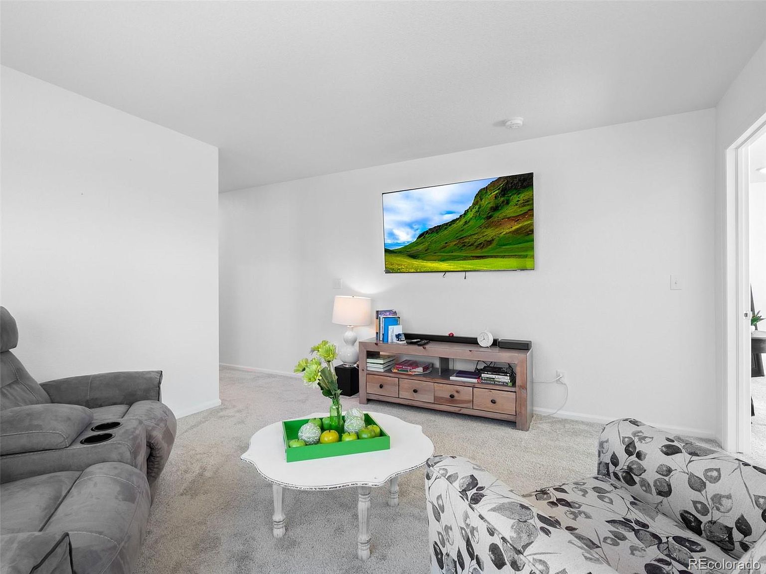 This is a bright and airy living room featuring neutral-toned walls and carpet. The room is furnished with a gray sofa, a floral armchair, and a light wood media console beneath a wall-mounted television. A white coffee table with decorative accents adds a touch of elegance to the space.