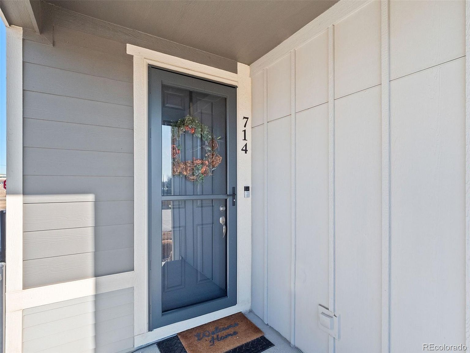 This image showcases the entryway of a home, featuring a gray door with a wreath, the house number '714', and a 'Welcome Home' doormat. The exterior is a combination of gray and white siding, creating a clean and inviting appearance. The perspective is a close-up, emphasizing the welcoming nature of the entrance.