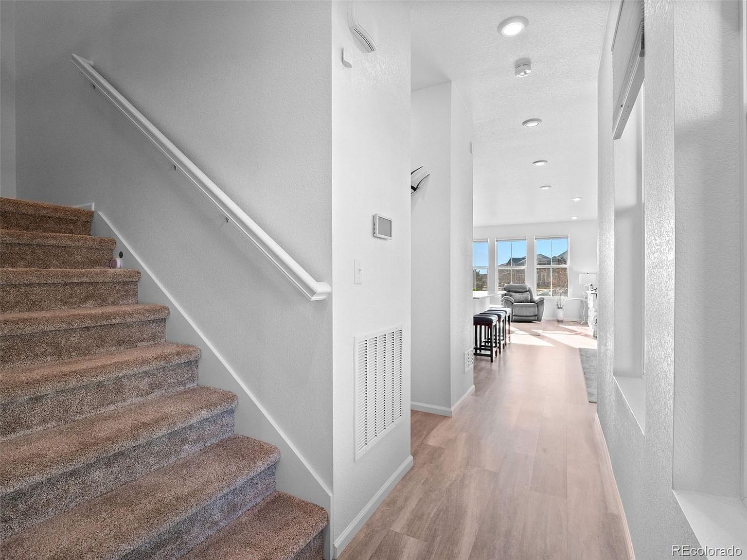 This interior shot showcases a bright hallway leading to a living area, with a staircase on the left. The hallway features light wood flooring and white walls, creating a clean and airy feel. The staircase is carpeted and has a white handrail. The perspective is from the entrance of the hallway, looking towards the living area.