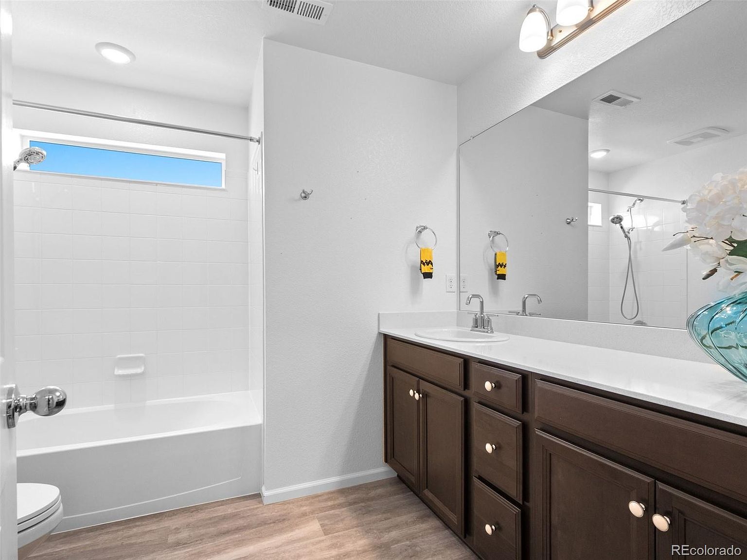 This is a well-lit bathroom featuring a white tub and shower combination with a clear window above the shower. The vanity has dark wood cabinets, a white countertop, and a single sink. A large mirror hangs above the vanity, reflecting the shower and adding to the sense of space.