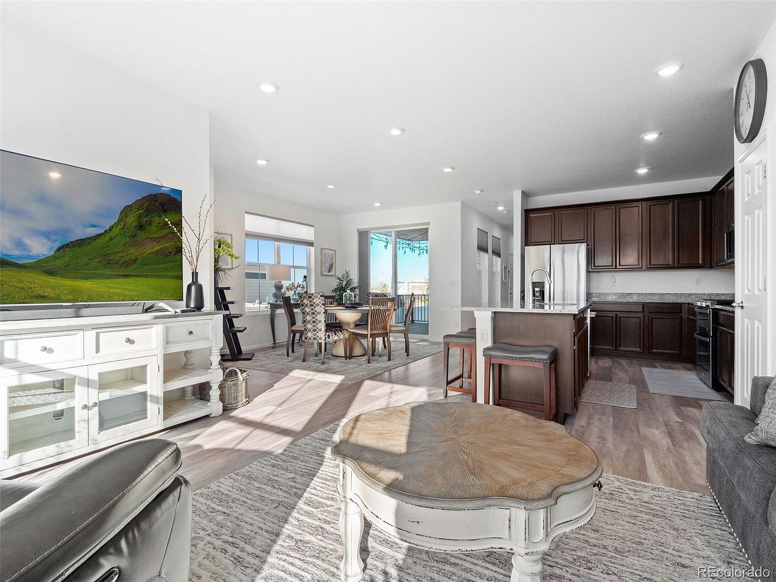 This is an interior shot of a living room that flows into a dining area and kitchen. The living room features a large TV, a white media console, a round coffee table, and a gray sofa. The dining area has a round table with chairs, and the kitchen has dark wood cabinets and stainless steel appliances. The overall impression is a bright, open, and modern living space.
