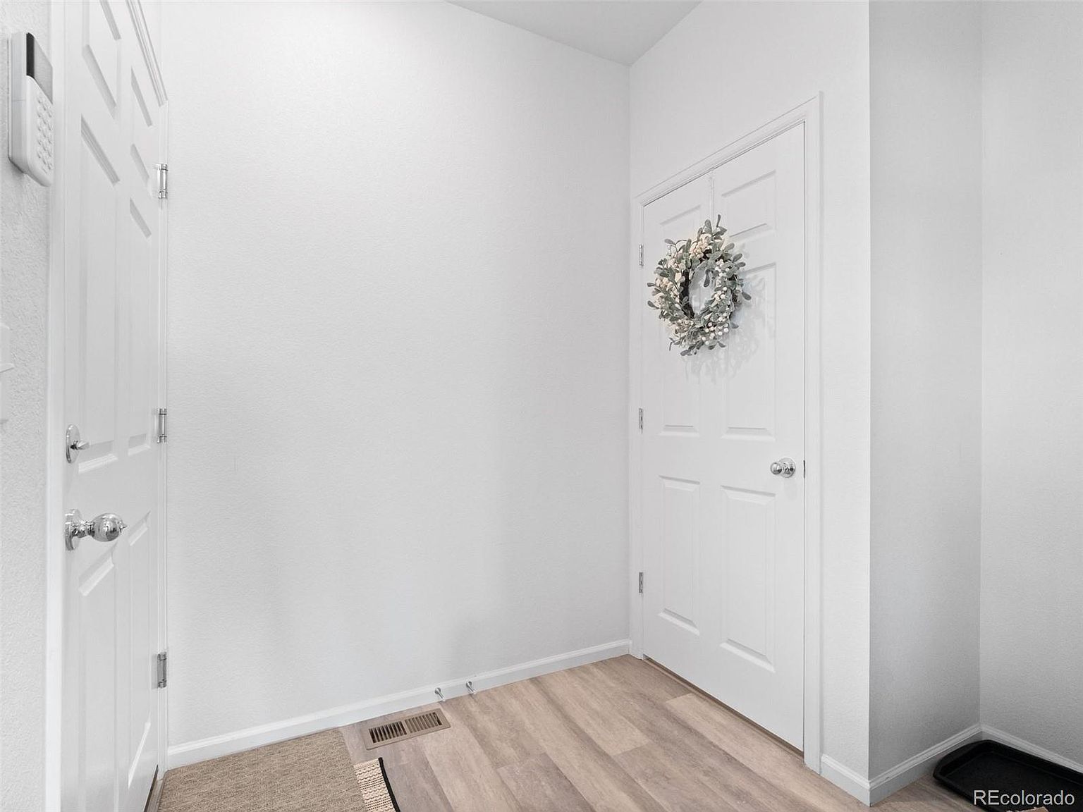 This is an interior shot of a hallway, likely an entryway. The walls are painted white, and the flooring is a light wood-look laminate. There are two white doors, one with a wreath, and a small rug near the entrance, creating a clean and welcoming atmosphere.