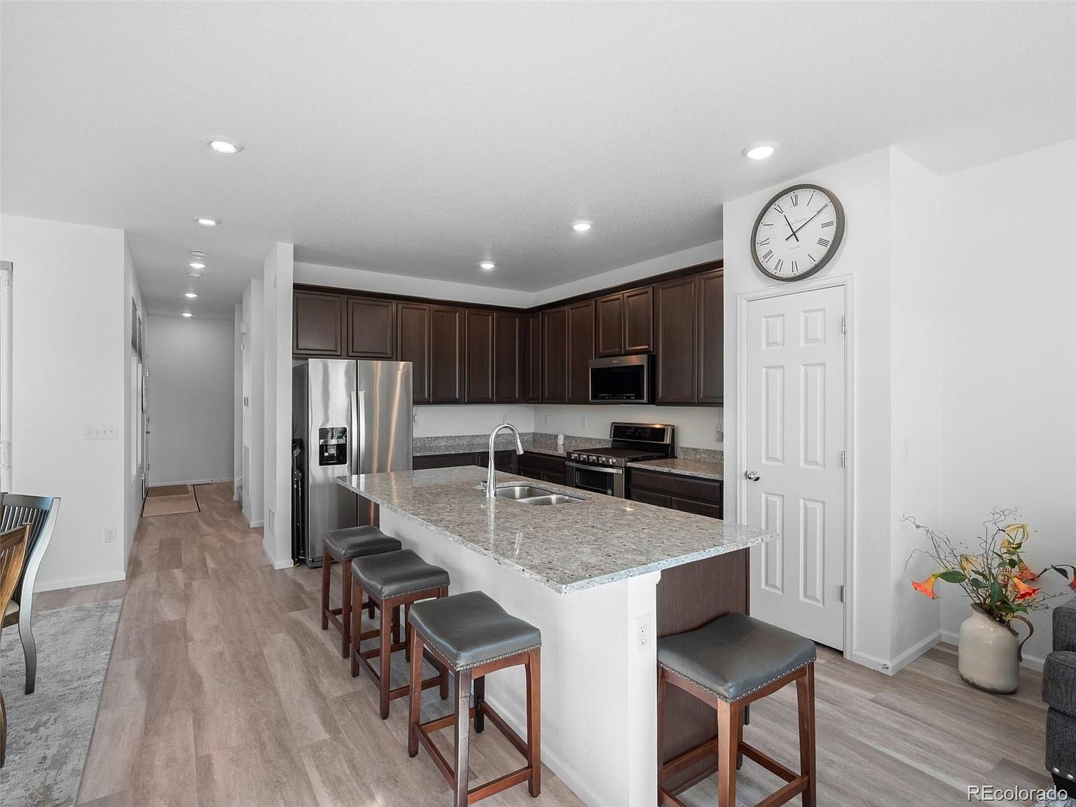 This is a well-lit kitchen featuring dark wood cabinetry, stainless steel appliances, and a granite countertop island with bar stool seating. The kitchen has a modern feel with recessed lighting and a clean, organized appearance. A large clock hangs on the wall near a white door, adding a touch of traditional charm.