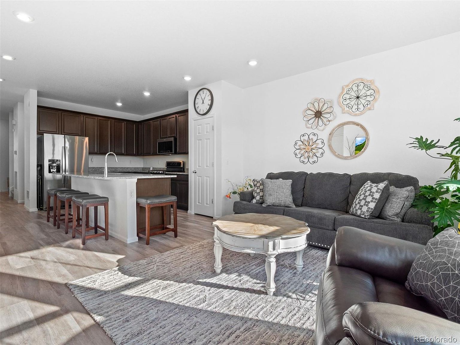 This is an interior shot of a living room that seamlessly blends into a kitchen area. The living room features a gray sofa with decorative pillows, a round coffee table, and a patterned rug. The kitchen area includes dark wood cabinets, stainless steel appliances, and a breakfast bar with stools, creating an open and inviting space.