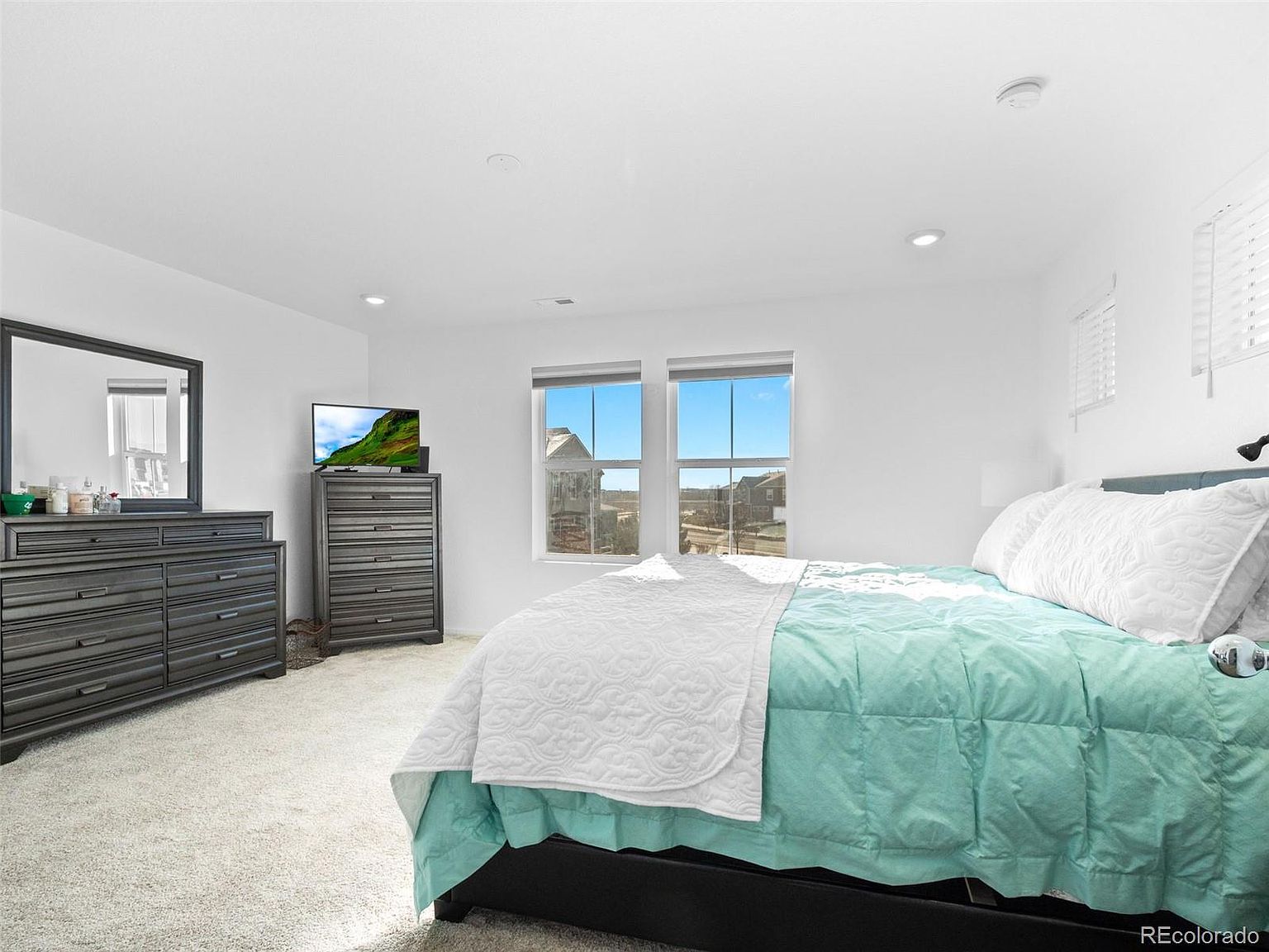This is a bright and airy primary bedroom featuring a large bed with a teal comforter and white quilt, two windows providing natural light, and a dresser with a mirror. A television sits atop a tall chest of drawers. The room is painted in a neutral color, creating a serene and inviting atmosphere.