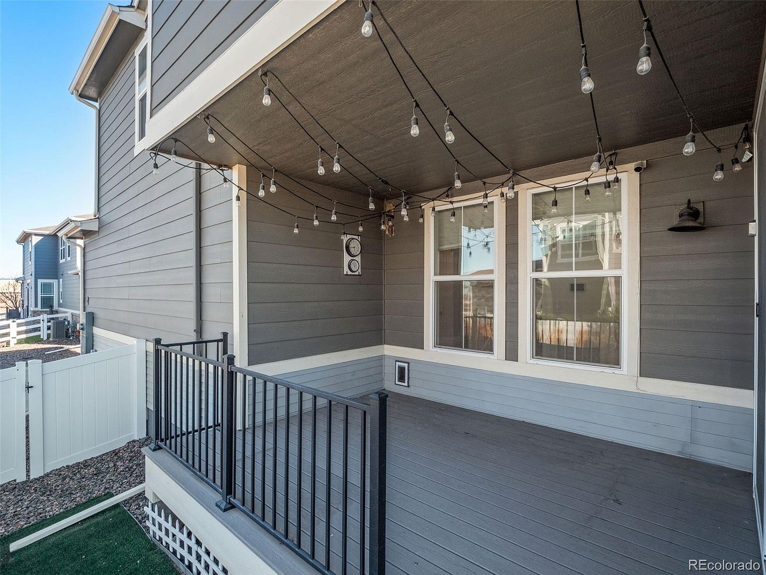 This image showcases a covered patio or deck area attached to a house. The space features a gray-toned deck, a black metal railing, and string lights hanging from the ceiling, creating a cozy and inviting atmosphere. The exterior of the house is clad in gray siding, and windows provide natural light to the interior.